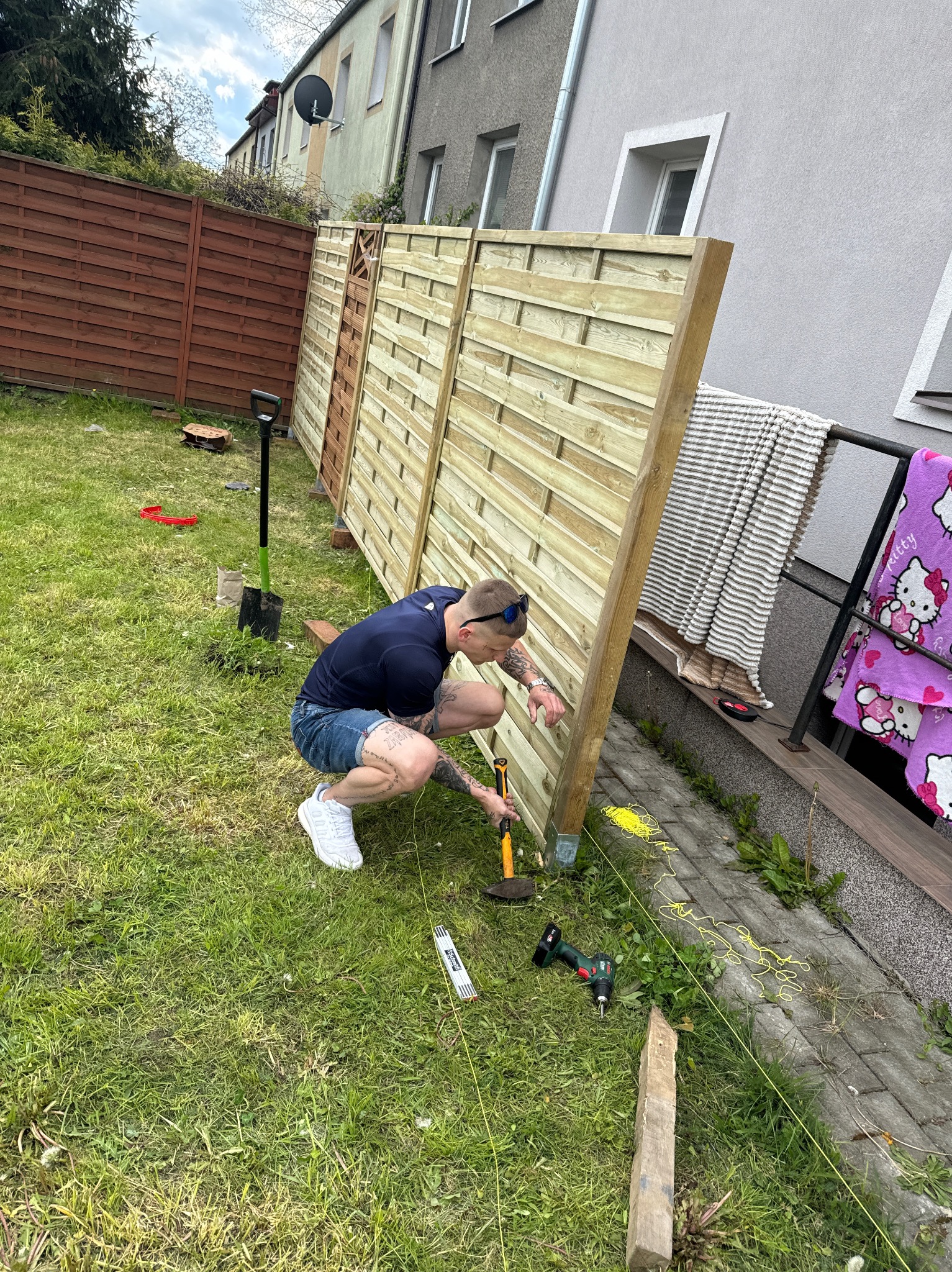 Mężczyzna w dżinsowych szortach i białych butach, z tatuażami na nogach i rękach, przykuca przy nowym drewnianym płocie, wyrównując go za pomocą młotka i poziomicy. W tle trawnik, budynek...