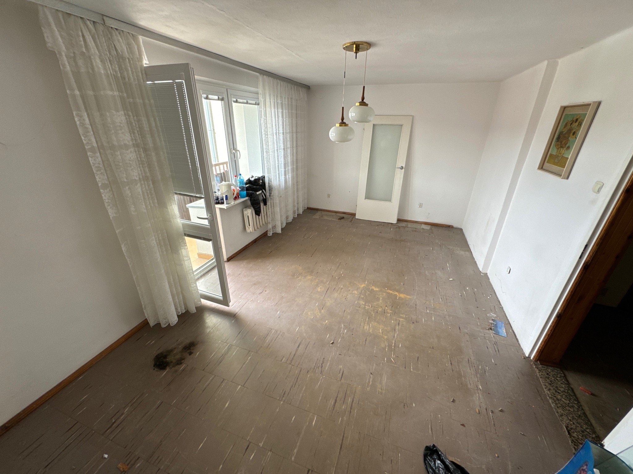 Pusty pokój w mieszkaniu po usunięciu mebli, z otwartym balkonem, odsłoniętymi podłogami z linoleum z widocznymi śladami użytkowania, lampą wiszącą w stylu retro i obrazem na ścianie.