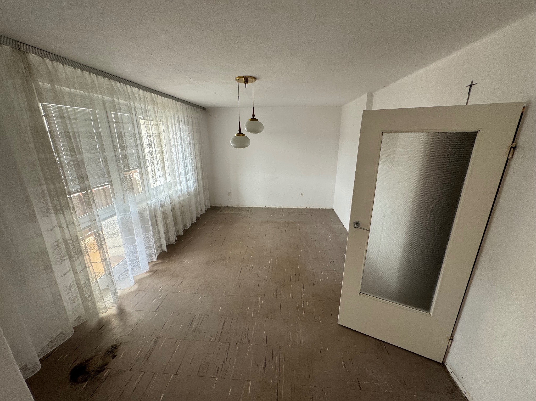 Pusty pokój przed remontem z brązową podłogą, białymi ścianami, oknem z firanką i wiszącą lampą. W rogu stoi białe drzwi z szybą. Na ścianie widoczny symbol krzyża.