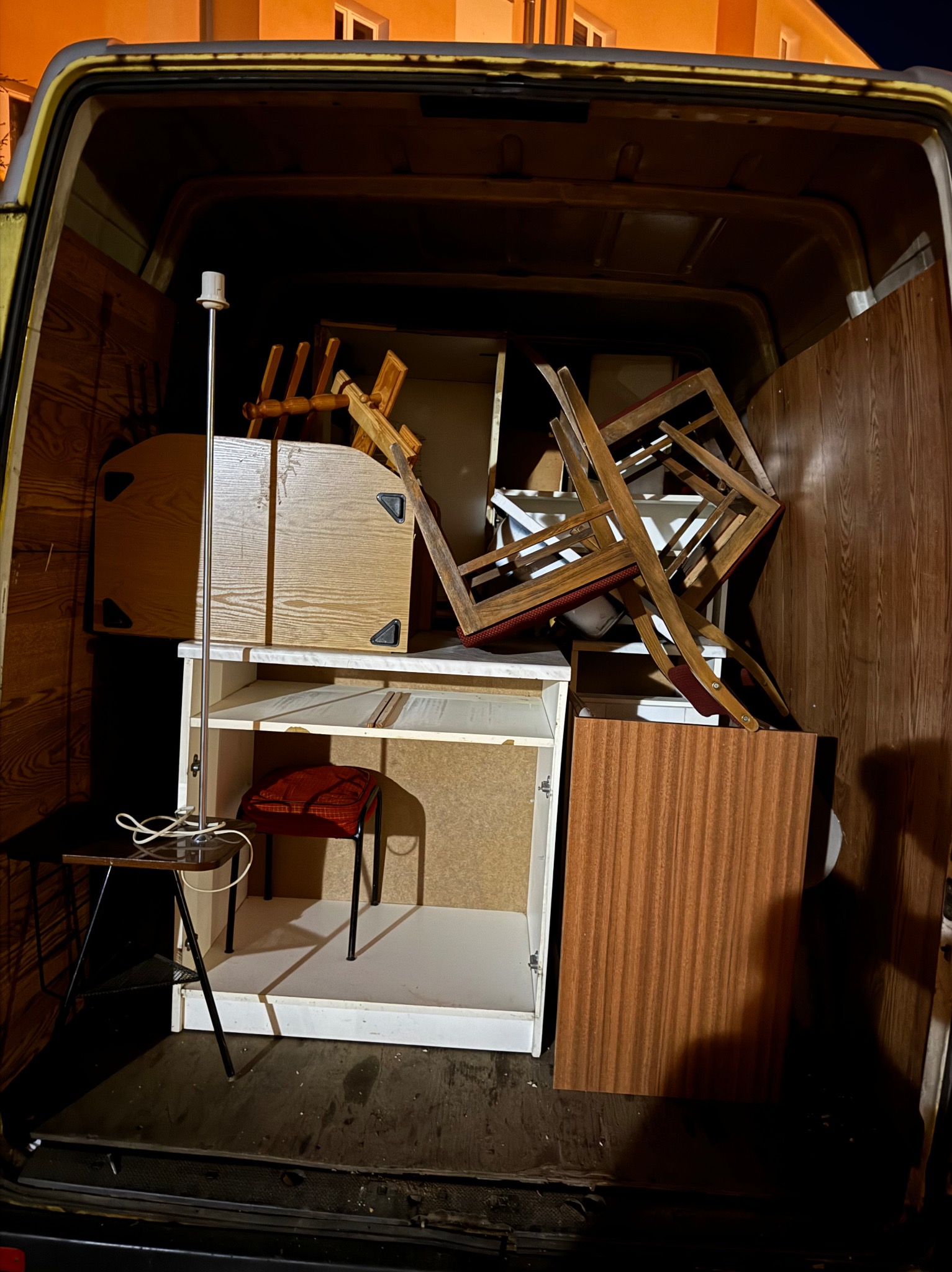 Wnętrze furgonetki załadowane meblami podczas przeprowadzki, widoczne krzesła, regał, stolik z lampą i inne przedmioty, oświetlenie sztuczne.