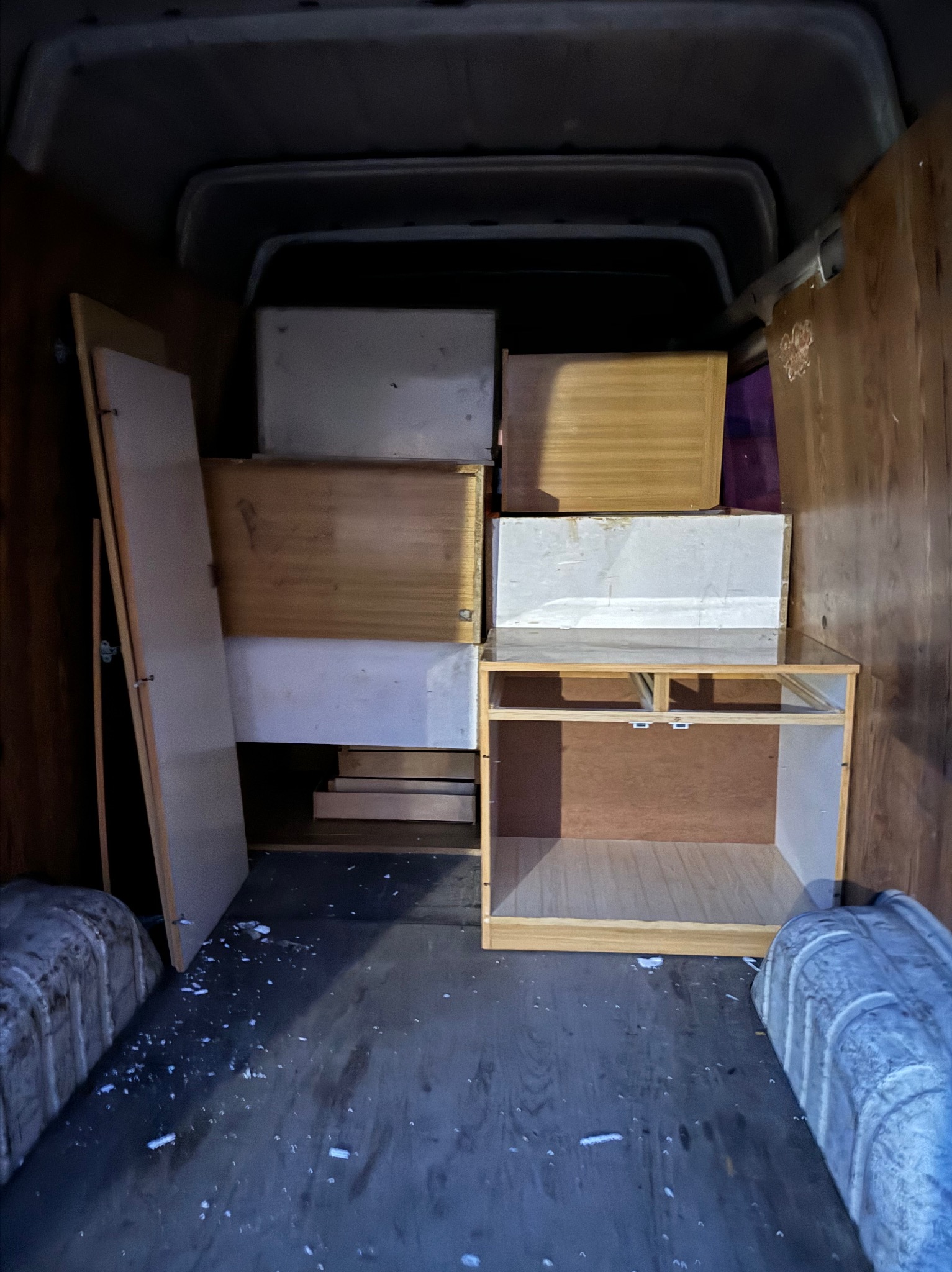 Wnętrze furgonetki załadowane meblami i kartonami podczas transportu, widoczne panele drewniane na ścianach i podłodze, z rozsypanymi białymi okruchami.