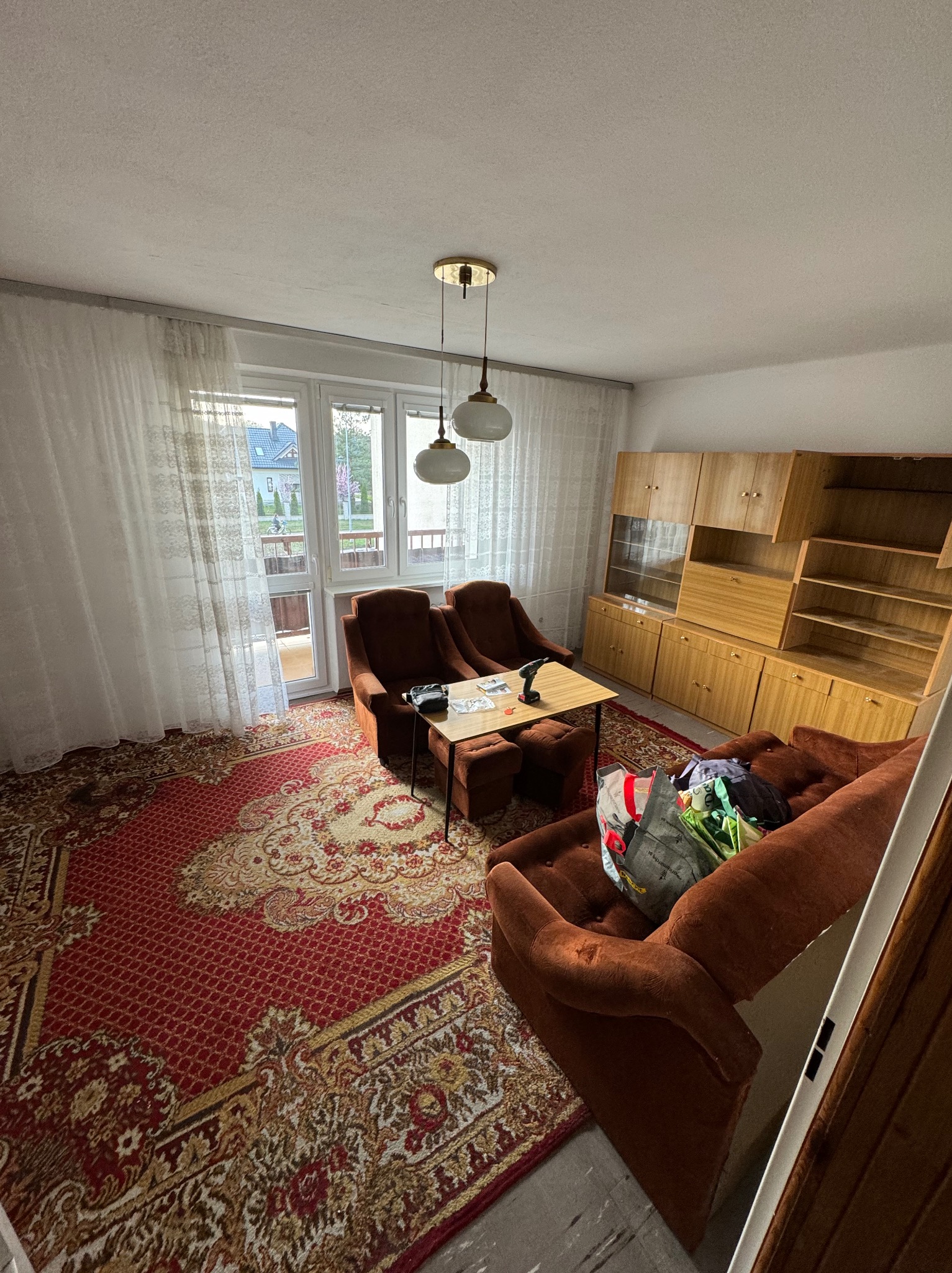 Pokój przed przeprowadzką: brązowe meble, dywan w orientalne wzory, kartony i torby spakowane na kanapie, wiertarka na stole, widok z balkonu.