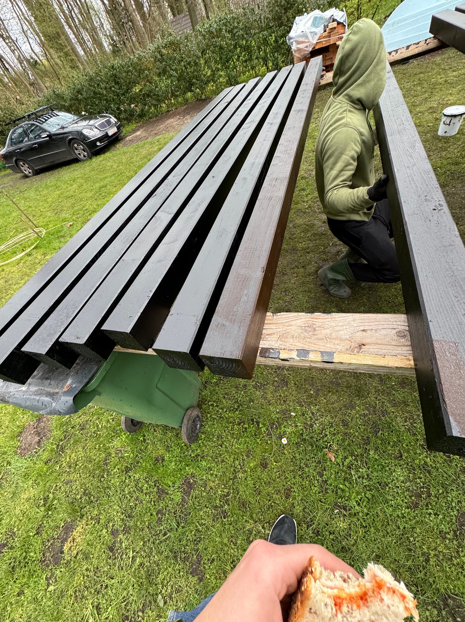 Malowanie drewnianych belek na czarno w plenerze, widok z góry na pracownika w zielonej bluzie z kapturem, obok czarny samochód i zielony kosz na śmieci, w kadrze dłoń z kanapką.
