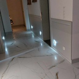 Best Project Sebastian Łobacz - Nowoczesny korytarz z białymi marmurowymi płytkami podłogowymi, panelami ściennymi z dyskretnym oświetleniem LED i wiszącymi obrazami w złotych ramach.