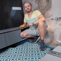 Best Project Sebastian Łobacz - Mężczyzna w trakcie montażu szafki łazienkowej, z miarką w dłoni, na tle marmurowych płytek i dywanika z geometrycznym wzorem.
