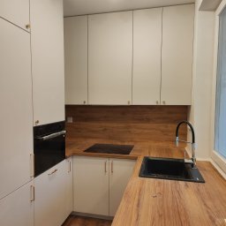 Best Project Sebastian Łobacz - Nowoczesna kuchnia z białymi szafkami i blatem imitującym drewno, czarny zlew i płyta indukcyjna, złote uchwyty, oświetlenie punktowe.