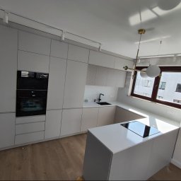 Best Project Sebastian Łobacz - Nowoczesna, minimalistyczna kuchnia w jasnych odcieniach szarości z wyspą kuchenną, czarną armaturą i oświetleniem punktowym oraz wiszącą lampą. Widok na zabudowę kuchenną z piekarnikiem i płytą...