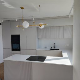 Best Project Sebastian Łobacz - Nowoczesna kuchnia z wyspą, szare fronty szafek z ryflowanym wzorem, biały blat, czarna płyta indukcyjna, złota lampa sufitowa z białymi kulami, oświetlenie szynowe.