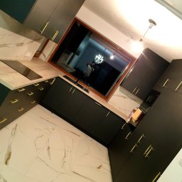 Best Project Sebastian Łobacz - Nowoczesna kuchnia z ciemnymi szafkami i złotymi uchwytami, wykończona płytkami imitującymi marmur, z dużym oknem odbijającym wnętrze i osobę robiącą zdjęcie.