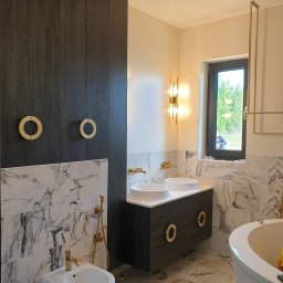 Best Project Sebastian Łobacz - Nowoczesna łazienka z marmurowymi płytkami, ciemną drewnianą zabudową i złotymi akcentami, w tym okrągłe uchwyty szafek i armatura, z widocznym bidetem i wanną.