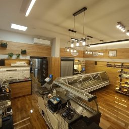 Best Project Sebastian Łobacz - Wnętrze sklepu spożywczego z drewnianymi elementami wykończenia, oświetleniem punktowym i wiszącymi lampami, widoczne lady chłodnicze, waga, kasy fiskalne oraz półki z produktami.