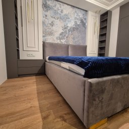 Best Project Sebastian Łobacz - Szare łóżko z weluru z niebieskim kocem w sypialni z zabudową w Rżyskach. W tle ściana z abstrakcyjnym wzorem i podłużne złote lampy.