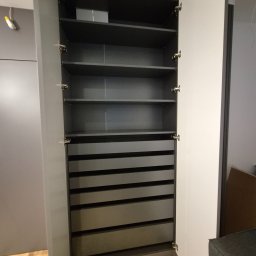 Best Project Sebastian Łobacz - Otwarta szafa wnękowa z szufladami i półkami w odcieniach szarości, widoczne zawiasy i drewniana podłoga.