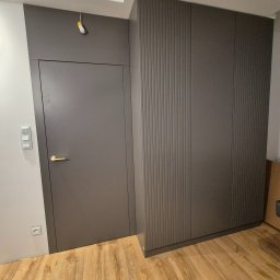 Best Project Sebastian Łobacz - Szara zabudowa ściany z ukrytymi drzwiami i szafą z pionowymi frezowaniami, złota klamka, drewniana podłoga, minimalistyczny styl.