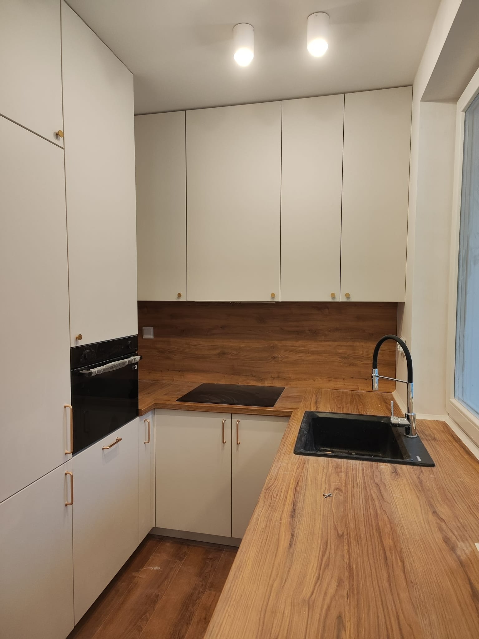 Nowoczesna kuchnia z białymi szafkami i blatem imitującym drewno, czarny zlew i płyta indukcyjna, złote uchwyty, oświetlenie punktowe.