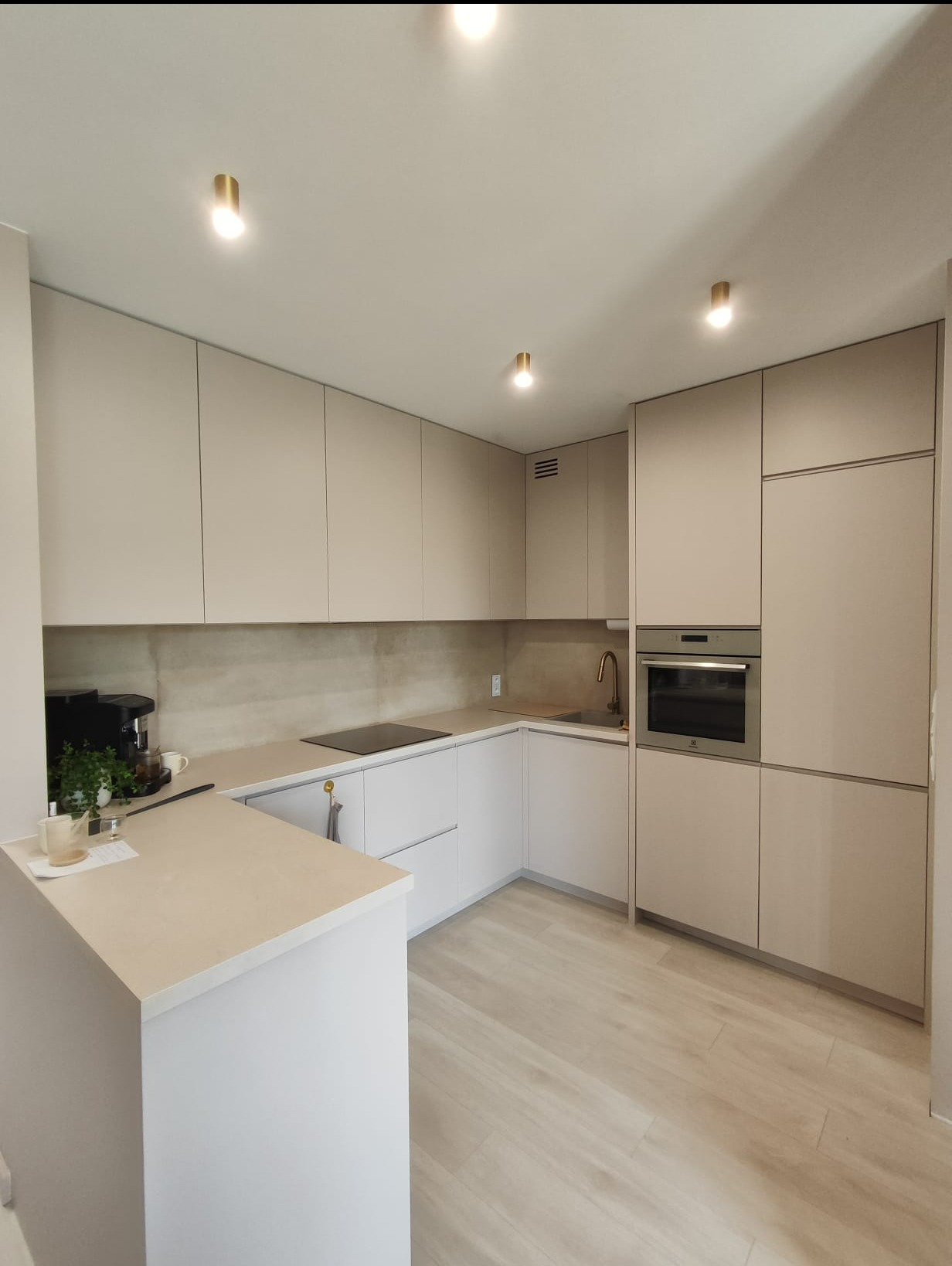 Nowoczesna kuchnia w odcieniach beżu i bieli z drewnopodobną podłogą, oświetleniem punktowym i minimalistycznym wyposażeniem, w tym ekspres do kawy i zlew z baterią.