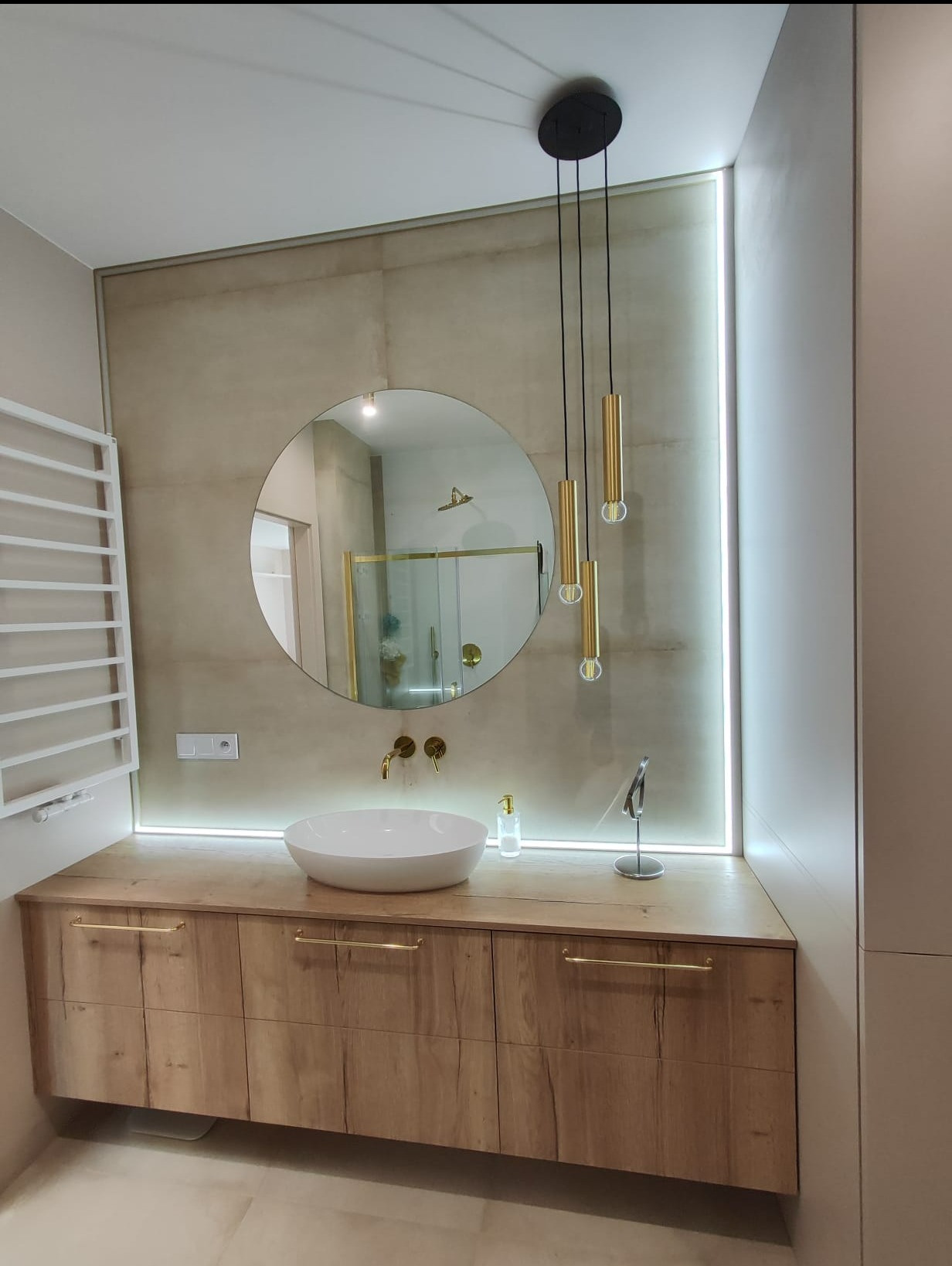 Nowoczesna łazienka z drewnianą szafką pod umywalką, okrągłym lustrem i złotymi akcentami, oświetlona listwą LED.