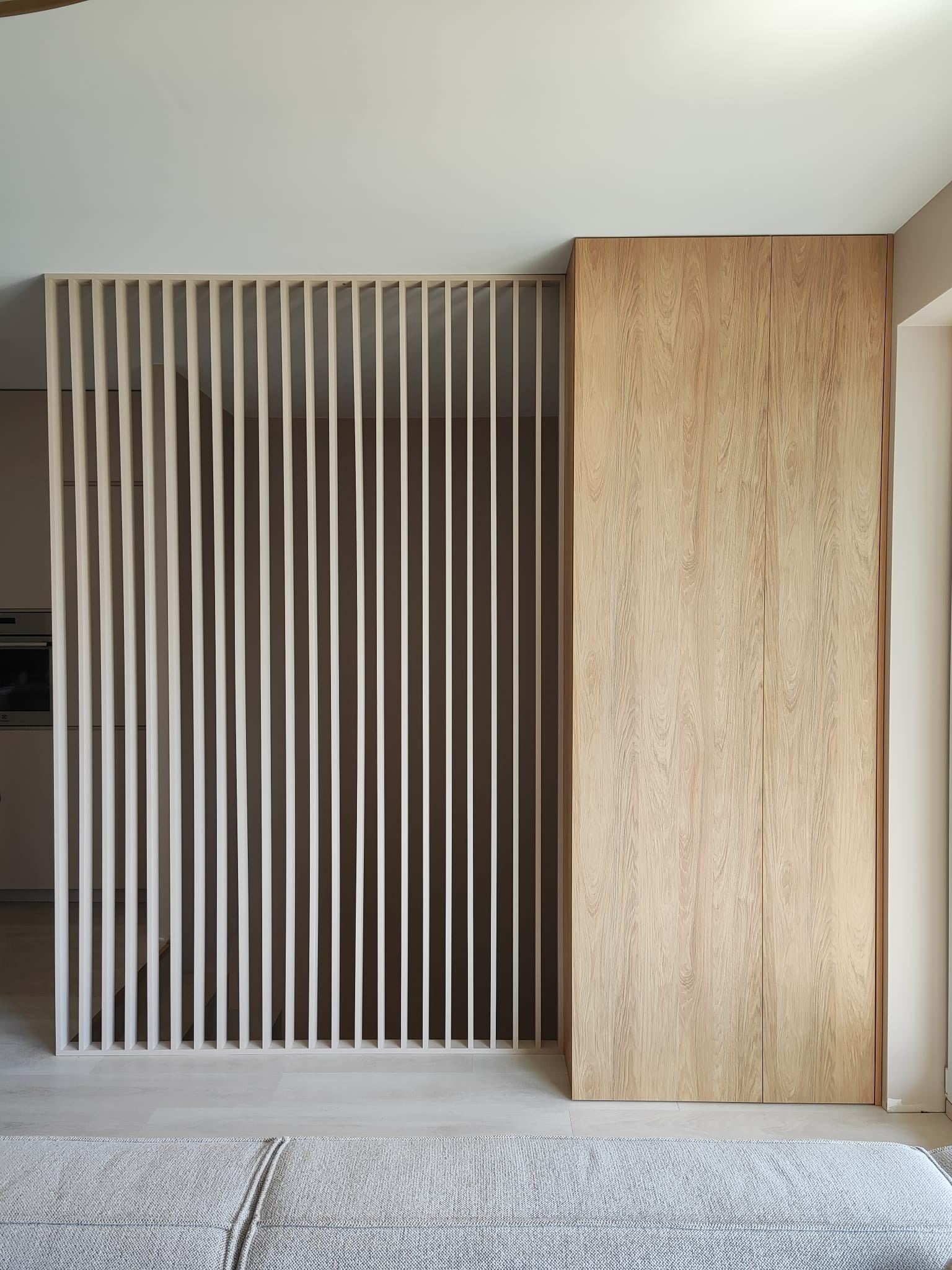 Minimalistyczna szafa wnękowa z jasnego drewna z przesuwnymi drzwiami, przylegająca do ażurowej ścianki działowej z pionowych listew w jasnym odcieniu, widoczna z perspektywy salonu.