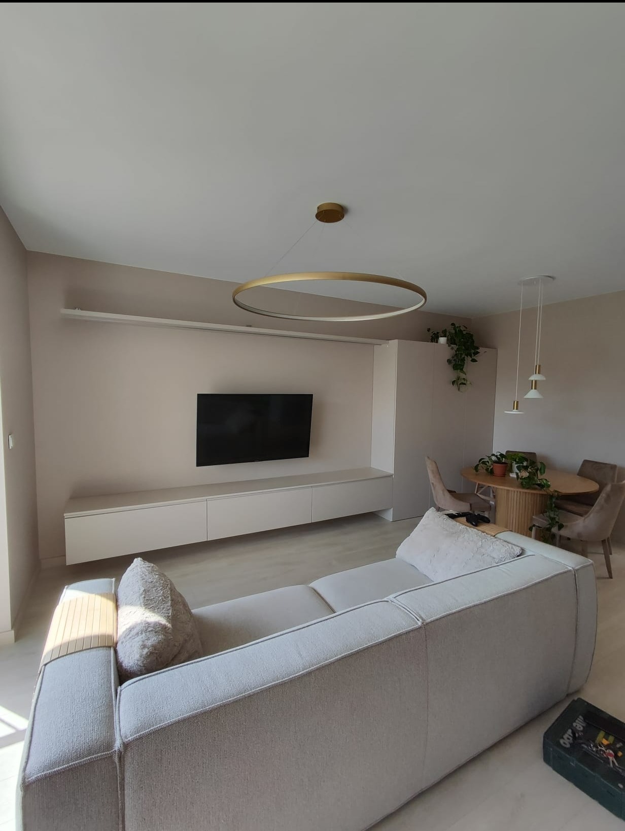 Jasne wnętrze salonu z szafką RTV pod telewizorem, okrągłym stołem jadalnym, sofą z poduszkami i okrągłą lampą sufitową. Minimalistyczny design z roślinami doniczkowymi.