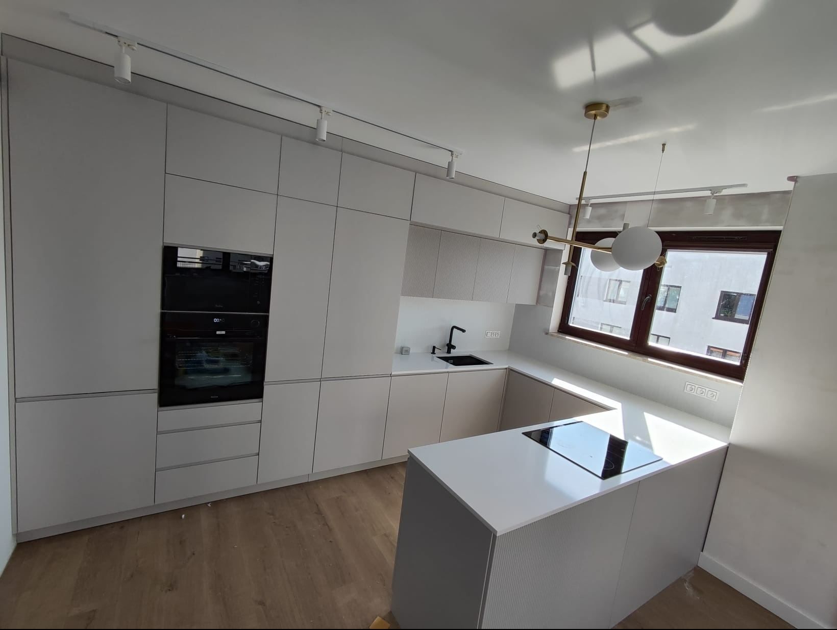 Nowoczesna, minimalistyczna kuchnia w jasnych odcieniach szarości z wyspą kuchenną, czarną armaturą i oświetleniem punktowym oraz wiszącą lampą. Widok na zabudowę kuchenną z piekarnikiem i płytą...