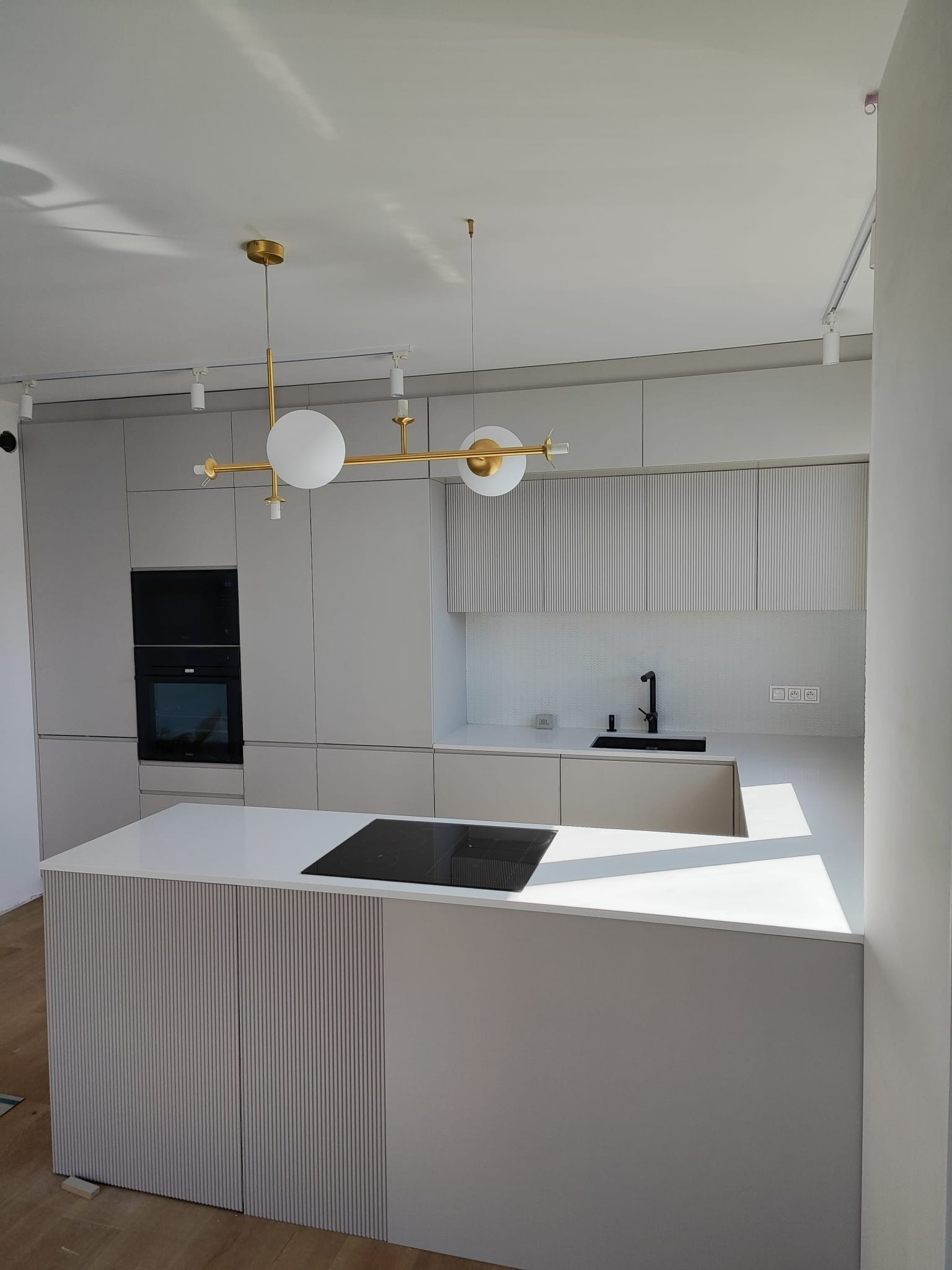 Nowoczesna kuchnia z wyspą, szare fronty szafek z ryflowanym wzorem, biały blat, czarna płyta indukcyjna, złota lampa sufitowa z białymi kulami, oświetlenie szynowe.