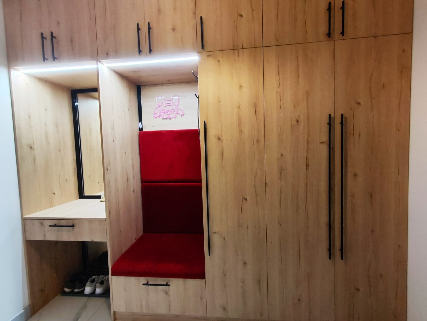 Zabudowa meblowa z drewnopodobnymi frontami, zawierająca szafę, siedzisko z czerwonym obiciem podświetlone neonem w kształcie pieska oraz toaletkę z lustrem i szufladą, uzupełniona o półkę na buty.