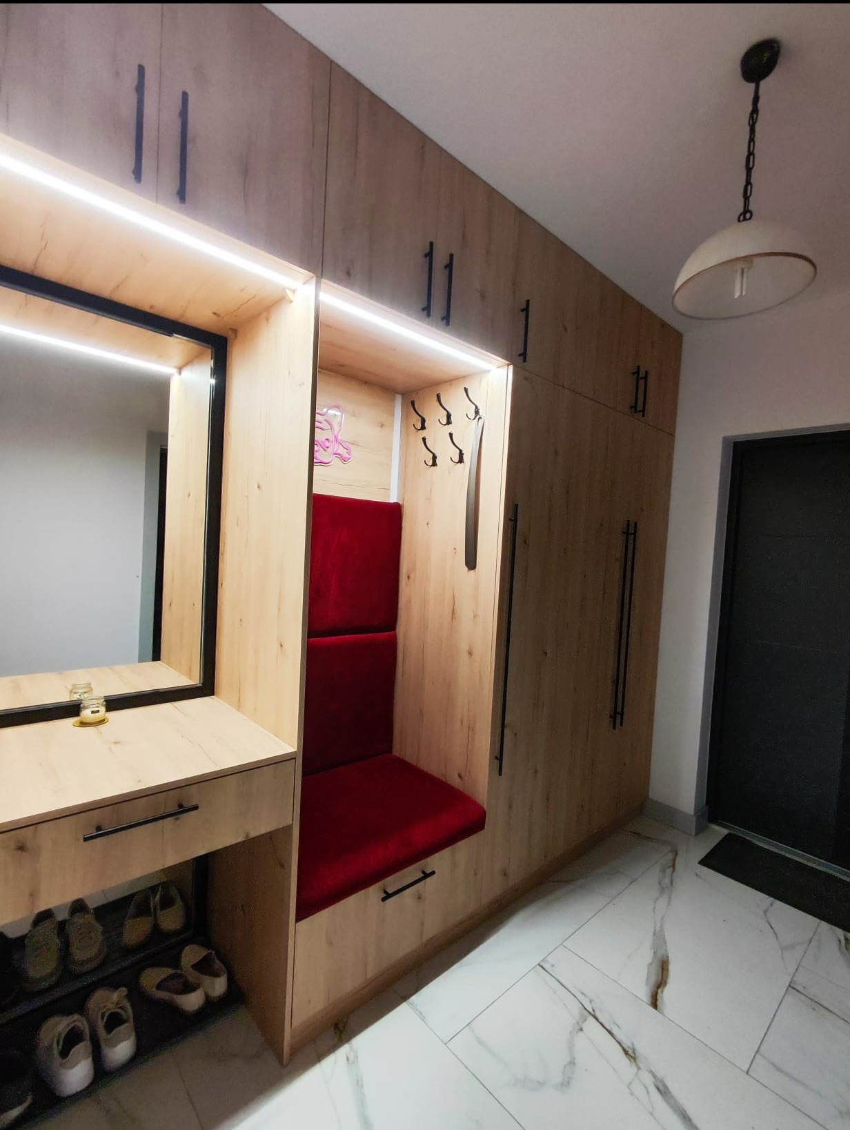 Zabudowa przedpokoju z siedziskiem obitym czerwoną tkaniną, lustrem z podświetleniem LED, wbudowanymi szafkami z długimi, czarnymi uchwytami, wieszakami na ubrania i półką na buty. Całość...