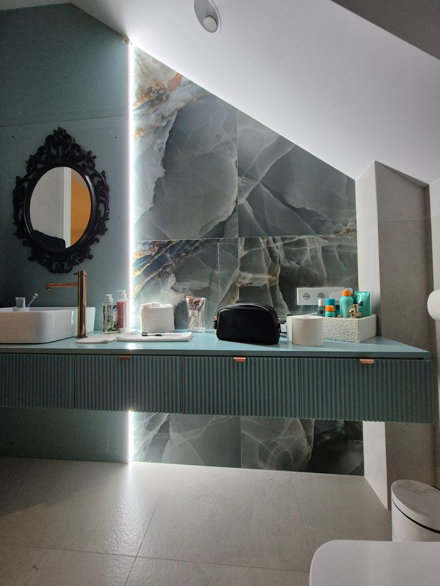 Nowoczesna łazienka na poddaszu z umywalką nablatową, baterią w kolorze złota, szafką z ryflowanym frontem w odcieniu turkusu i lustrem w ozdobnej, czarnej ramie. Ściana wykończona płytami...