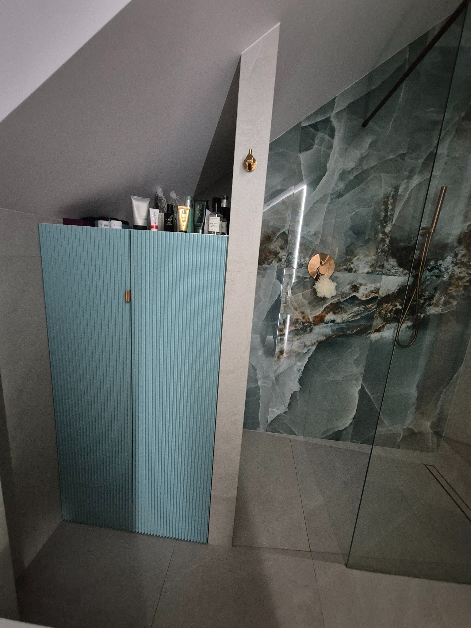 Niebieska szafka łazienkowa z ryflowanymi drzwiami, umieszczona pod skosem dachu, z kosmetykami na górze, obok kabina prysznicowa z marmurową ścianą.