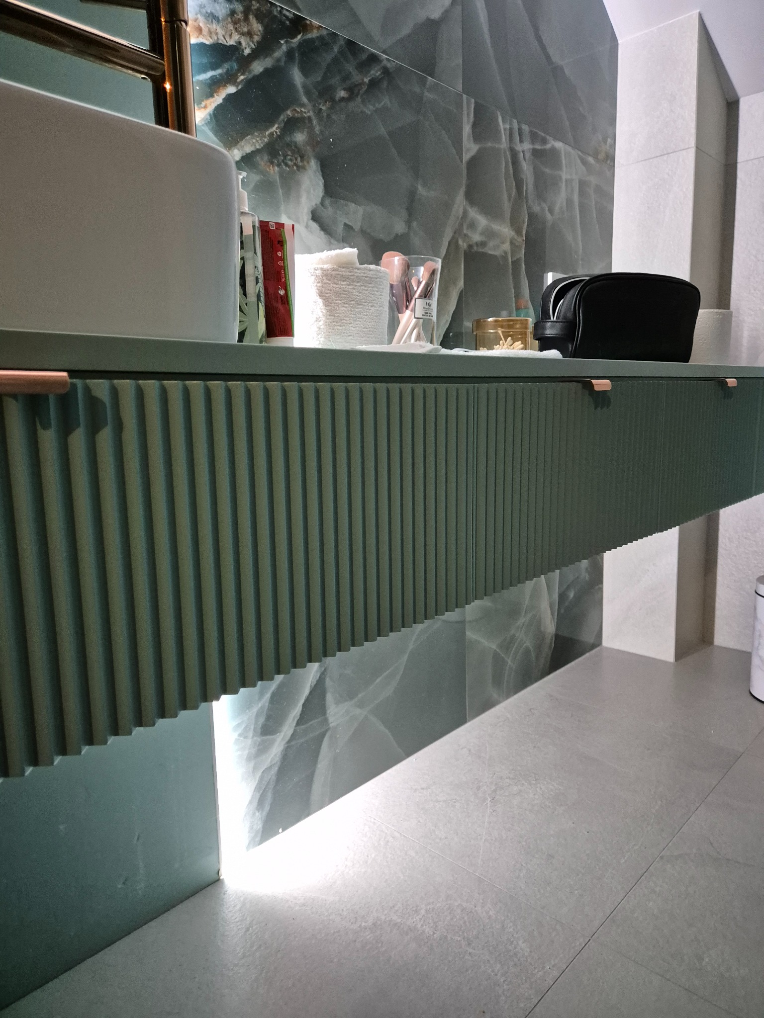 Nowoczesna łazienka z szafką pod umywalkę o ryflowanym froncie w odcieniach zieleni, marmurowa ściana w tle, oświetlenie LED pod szafką, kosmetyki i akcesoria na blacie.