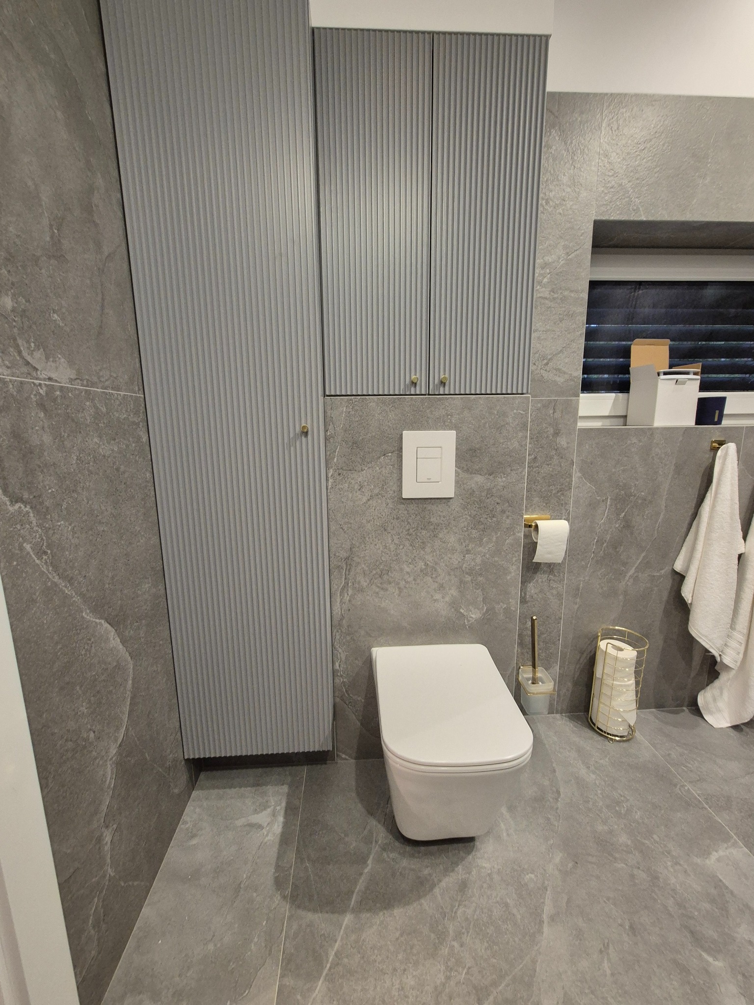 Nowoczesna toaleta podwieszana z szarą zabudową meblową z ryflowanymi frontami, dopasowaną kolorystycznie do płytek imitujących marmur na ścianach i podłodze.