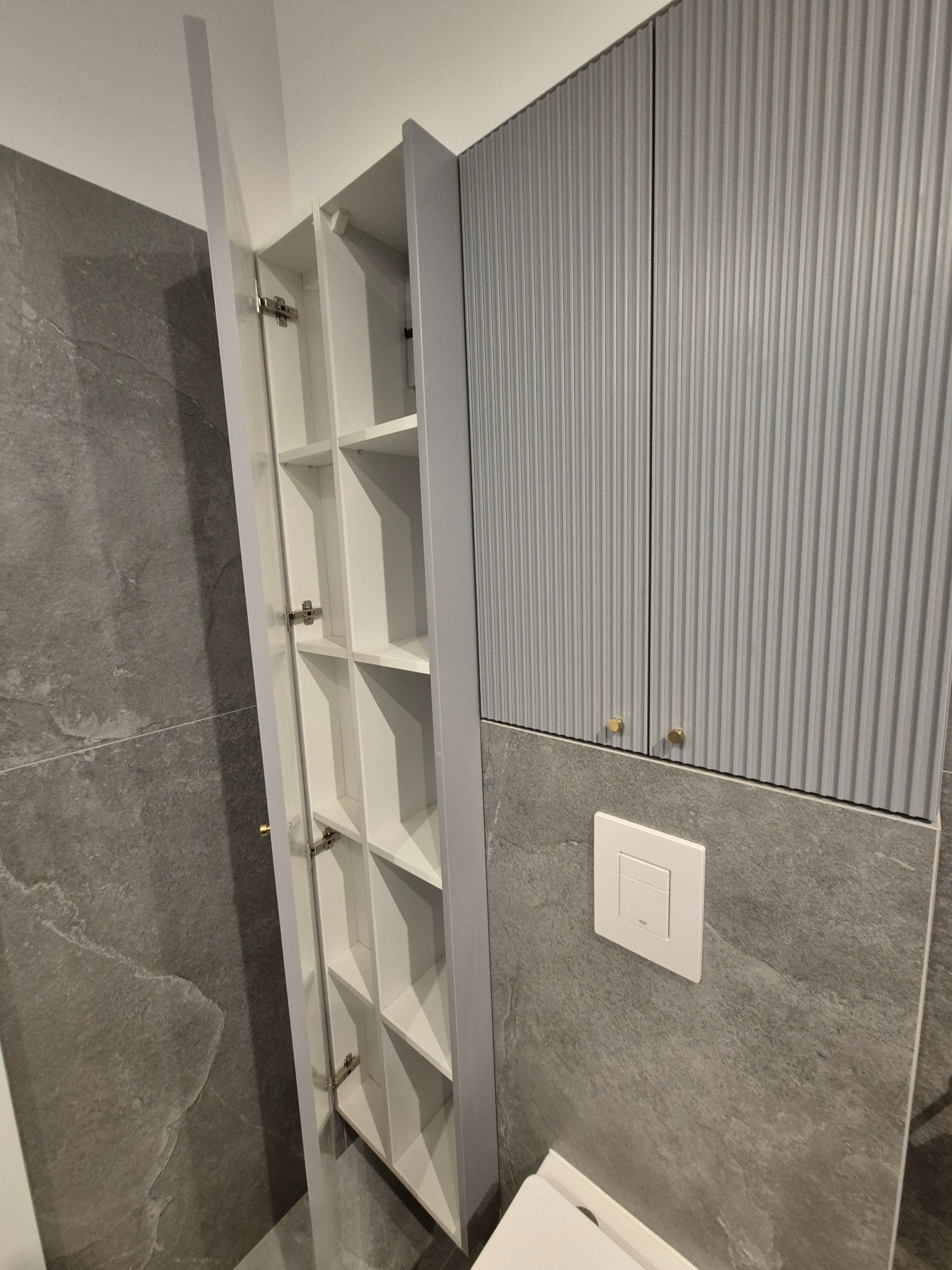 Wąska szafka łazienkowa z otwartymi drzwiami, ukazująca białe półki, obok szarej szafki z ryflowanymi frontami i złotymi gałkami, na tle szarych płytek imitujących kamień.