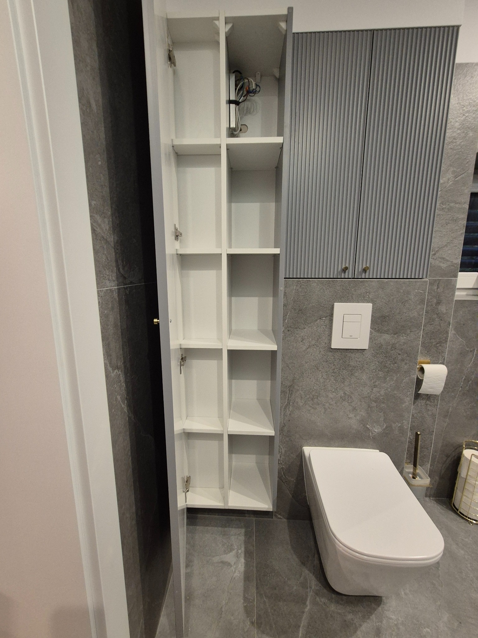 Wnętrze łazienki z białą, wysoką szafką z otwartymi półkami, szarą zabudową nad toaletą i podwieszaną miską WC na tle szarych płytek imitujących kamień.