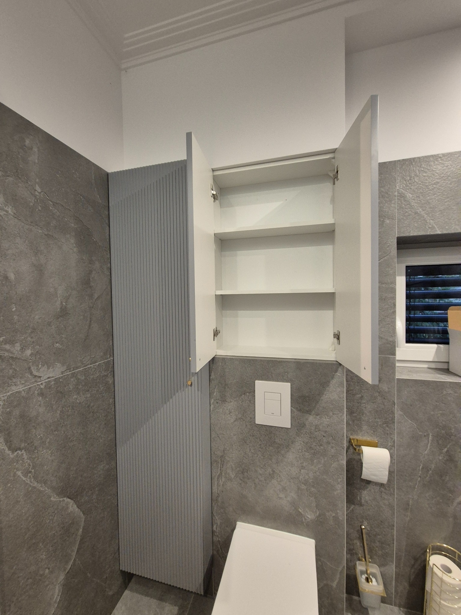 Szara łazienka z wiszącą szafką z otwartymi białymi drzwiczkami, widoczne półki wewnątrz, obok pionowa szafka z ryflowanym frontem, toaleta i uchwyt na papier toaletowy w złotym kolorze.
