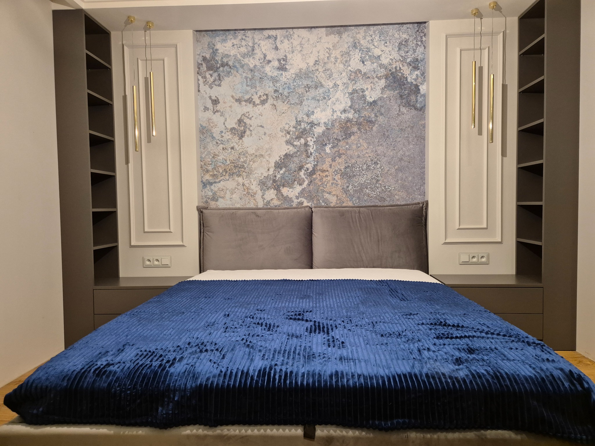 Nowoczesna sypialnia z łóżkiem z granatową narzutą, szarą tapetą imitującą marmur, szafkami wnękowymi po bokach i złotymi lampami wiszącymi.