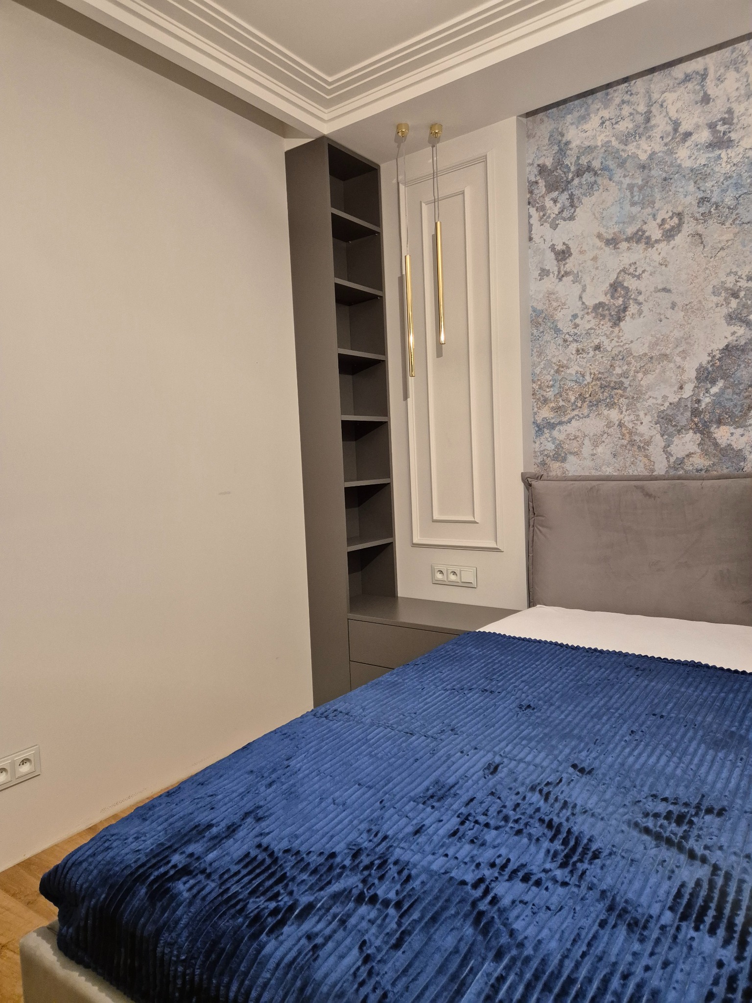 Nowoczesna sypialnia z szarą szafą wnękową z otwartymi półkami, zamontowaną obok łóżka z niebieską narzutą i szarym zagłówkiem, ściana z dekoracyjną tapetą w odcieniach błękitu i szarości.