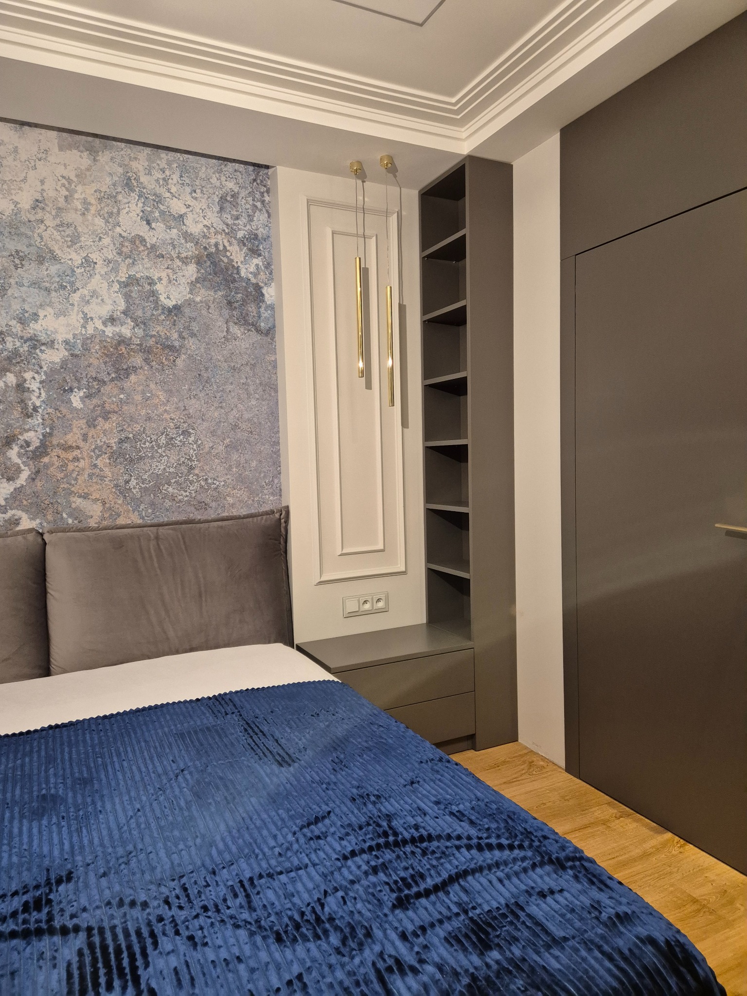 Nowoczesna sypialnia z szarą zabudową szafy wnękowej, otwartymi półkami, tapetą imitującą marmur, i dekoracyjnym oświetleniem wiszącym.