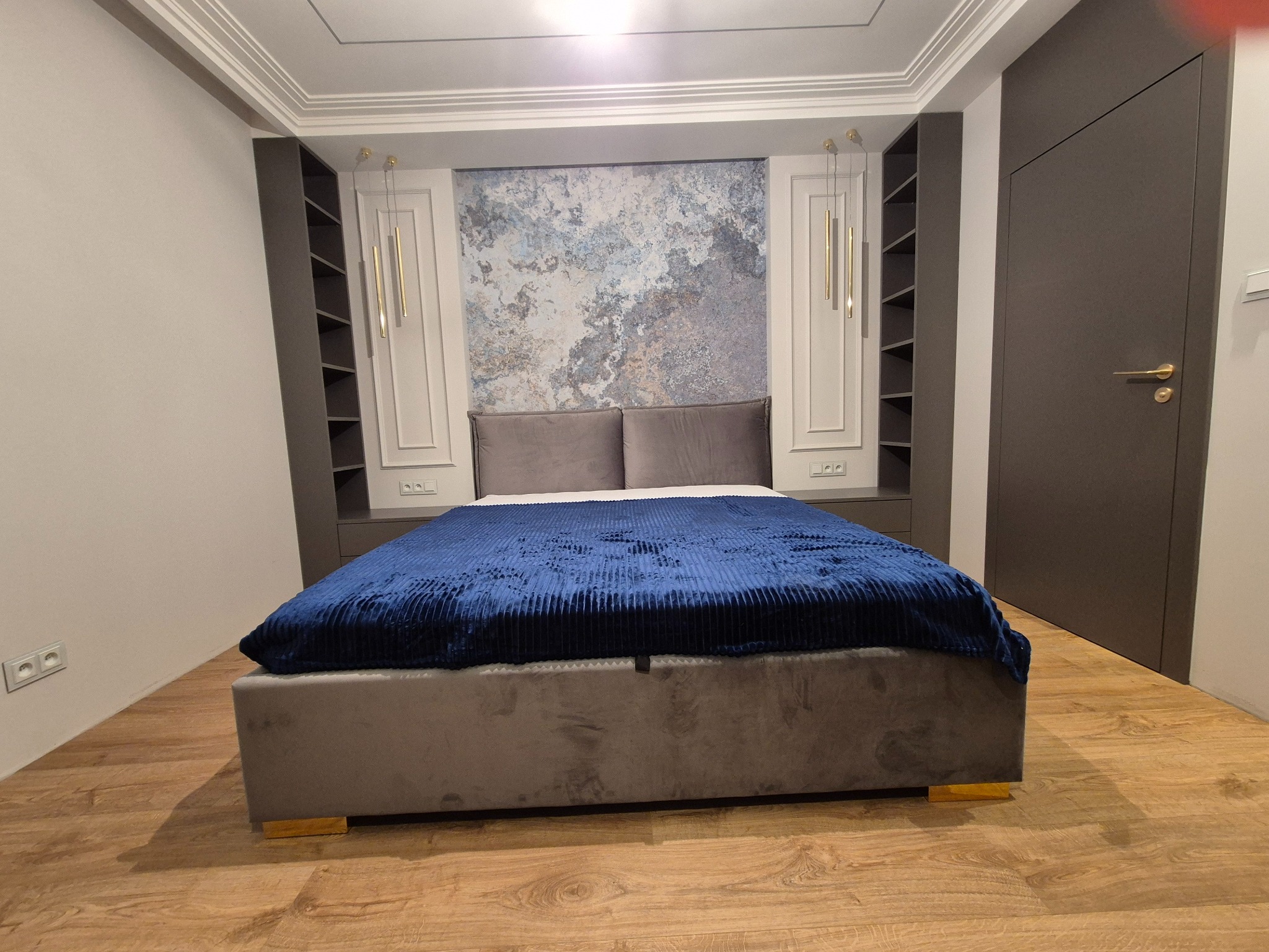 Nowoczesna sypialnia z łóżkiem kontynentalnym, niebieską narzutą, szarą tapetą imitującą marmur, symetrycznymi szafkami w zabudowie i złotymi lampami wiszącymi.