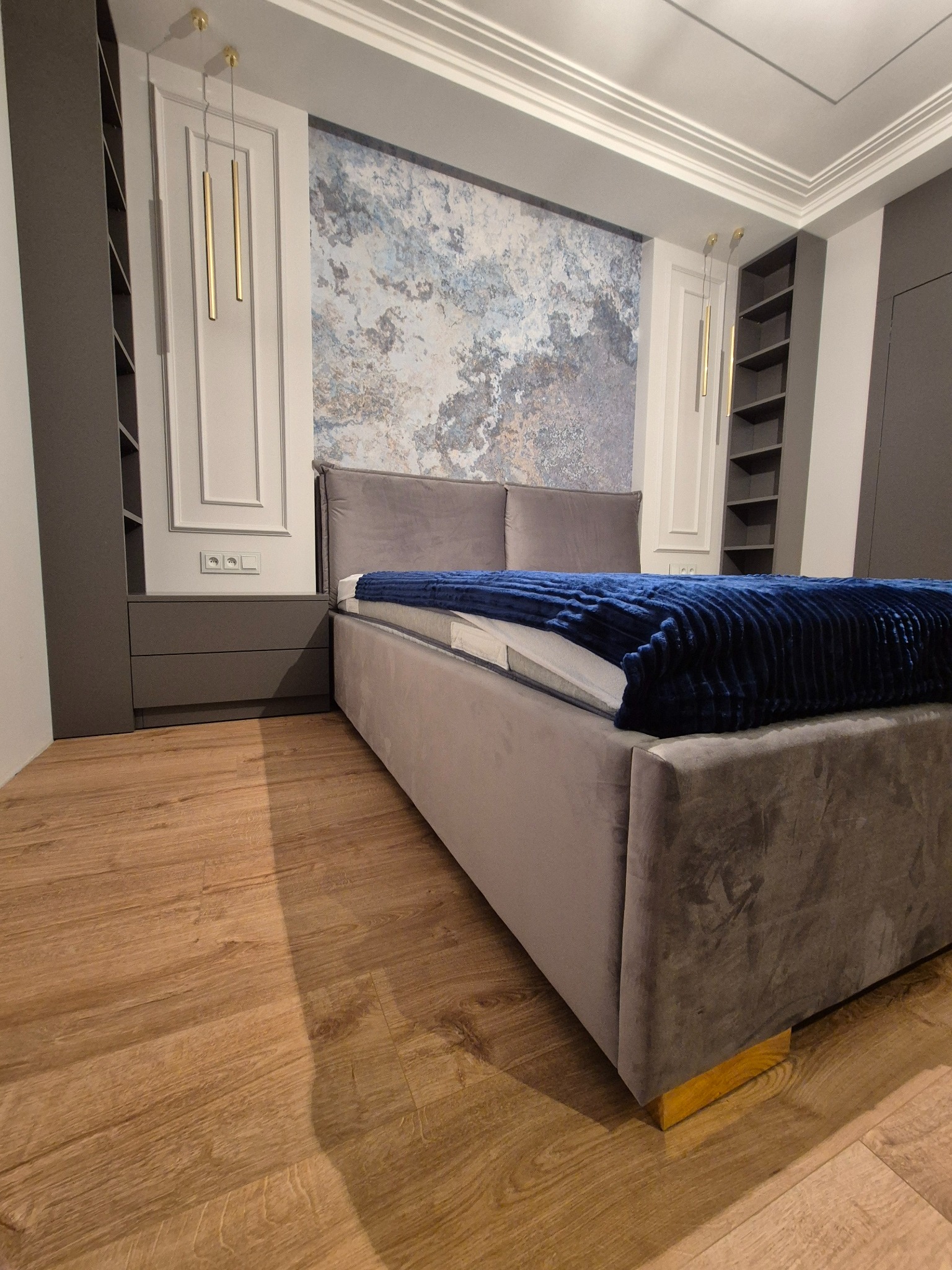 Szare łóżko z weluru z niebieskim kocem w sypialni z zabudową w Rżyskach. W tle ściana z abstrakcyjnym wzorem i podłużne złote lampy.