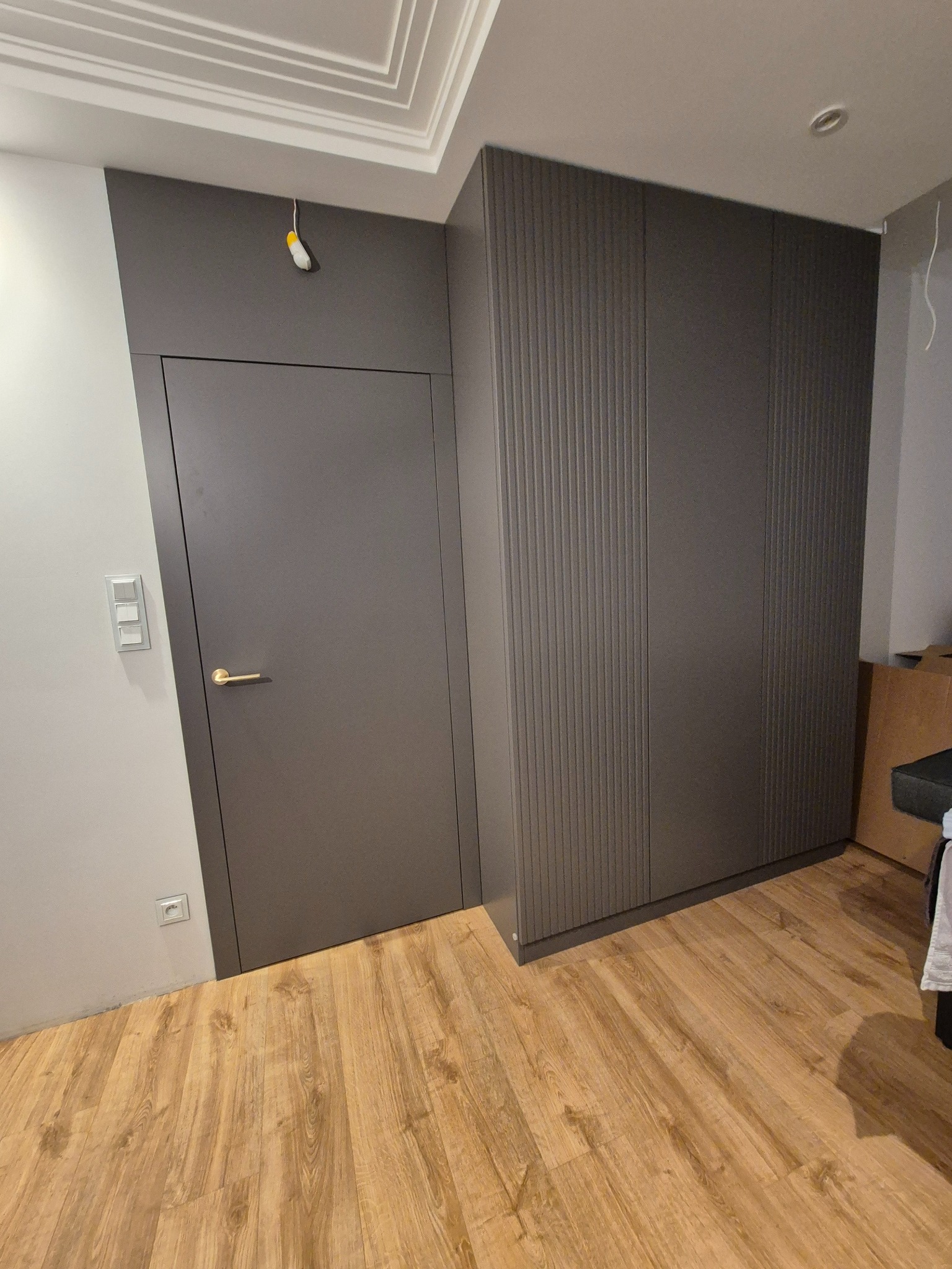 Grafitowa szafa wnękowa z ryflowanymi frontami, zintegrowana z drzwiami, na jasnej drewnianej podłodze; widoczny fragment sufitu z dekoracyjną sztukaterią.