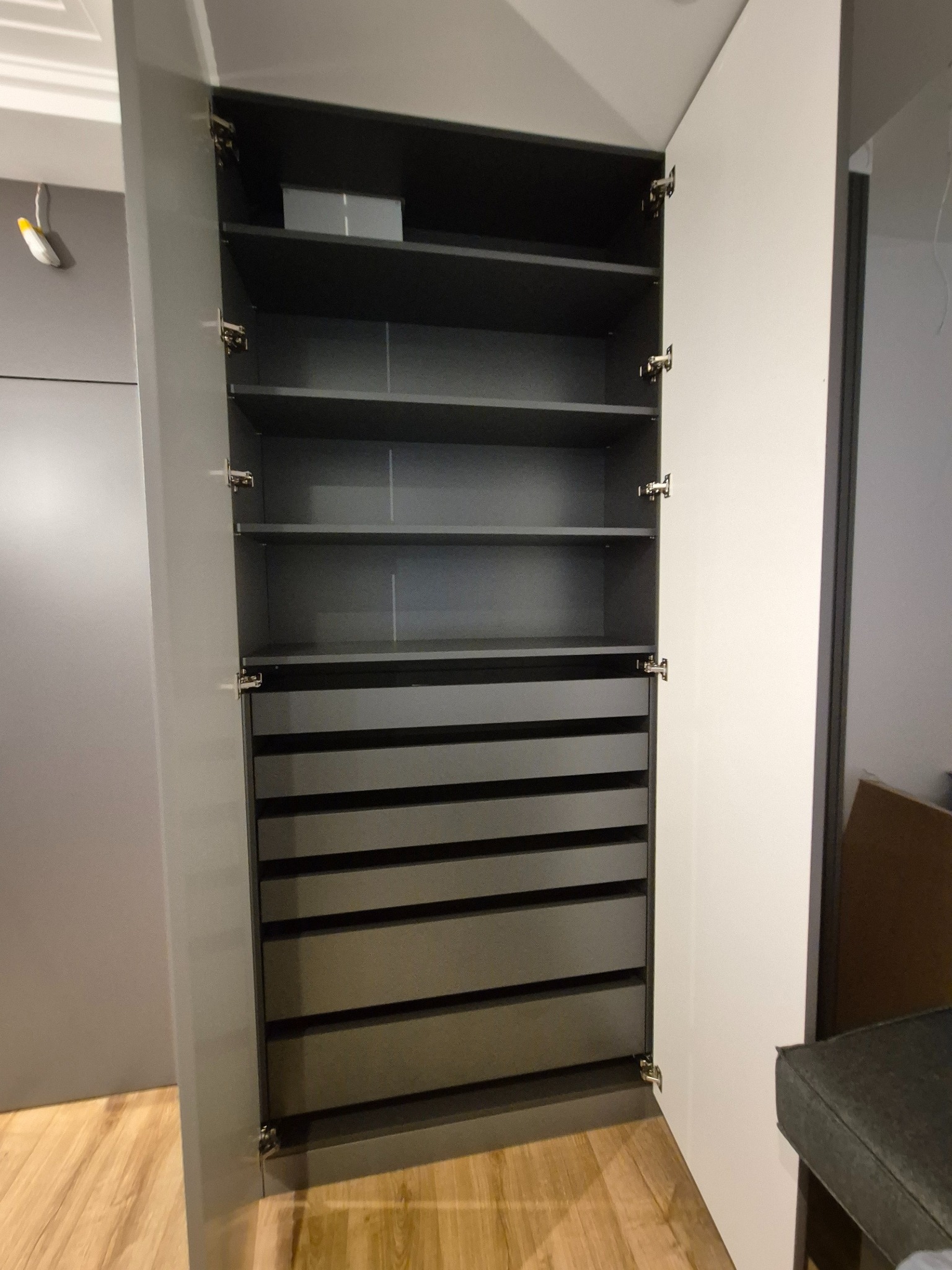 Otwarta szafa wnękowa z szufladami i półkami w odcieniach szarości, widoczne zawiasy i drewniana podłoga.