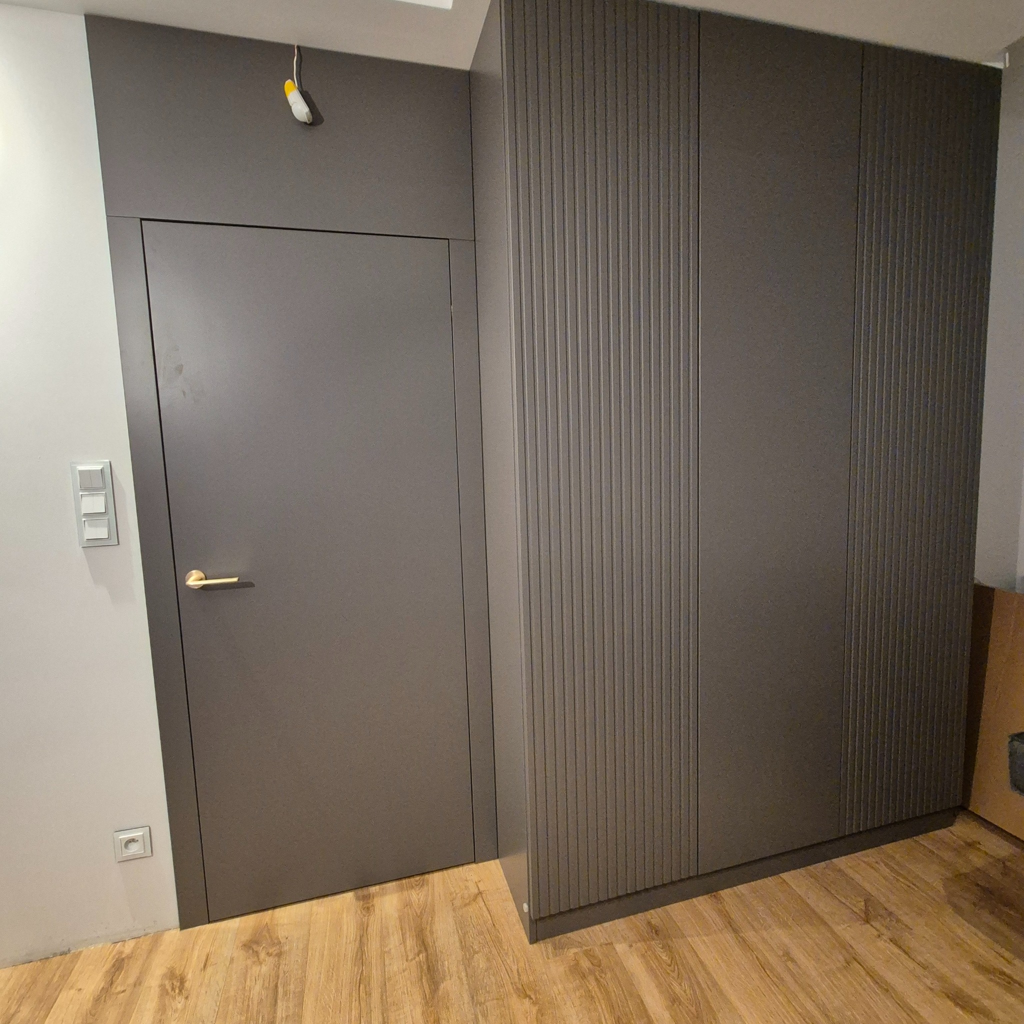 Szara zabudowa ściany z ukrytymi drzwiami i szafą z pionowymi frezowaniami, złota klamka, drewniana podłoga, minimalistyczny styl.