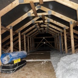 X-Term - Poddasze z drewnianą konstrukcją więźby dachowej, częściowo ocieplone białą wełną mineralną. Widoczny agregat do wdmuchiwania izolacji. Ujęcie z dołu, w głąb poddasza.