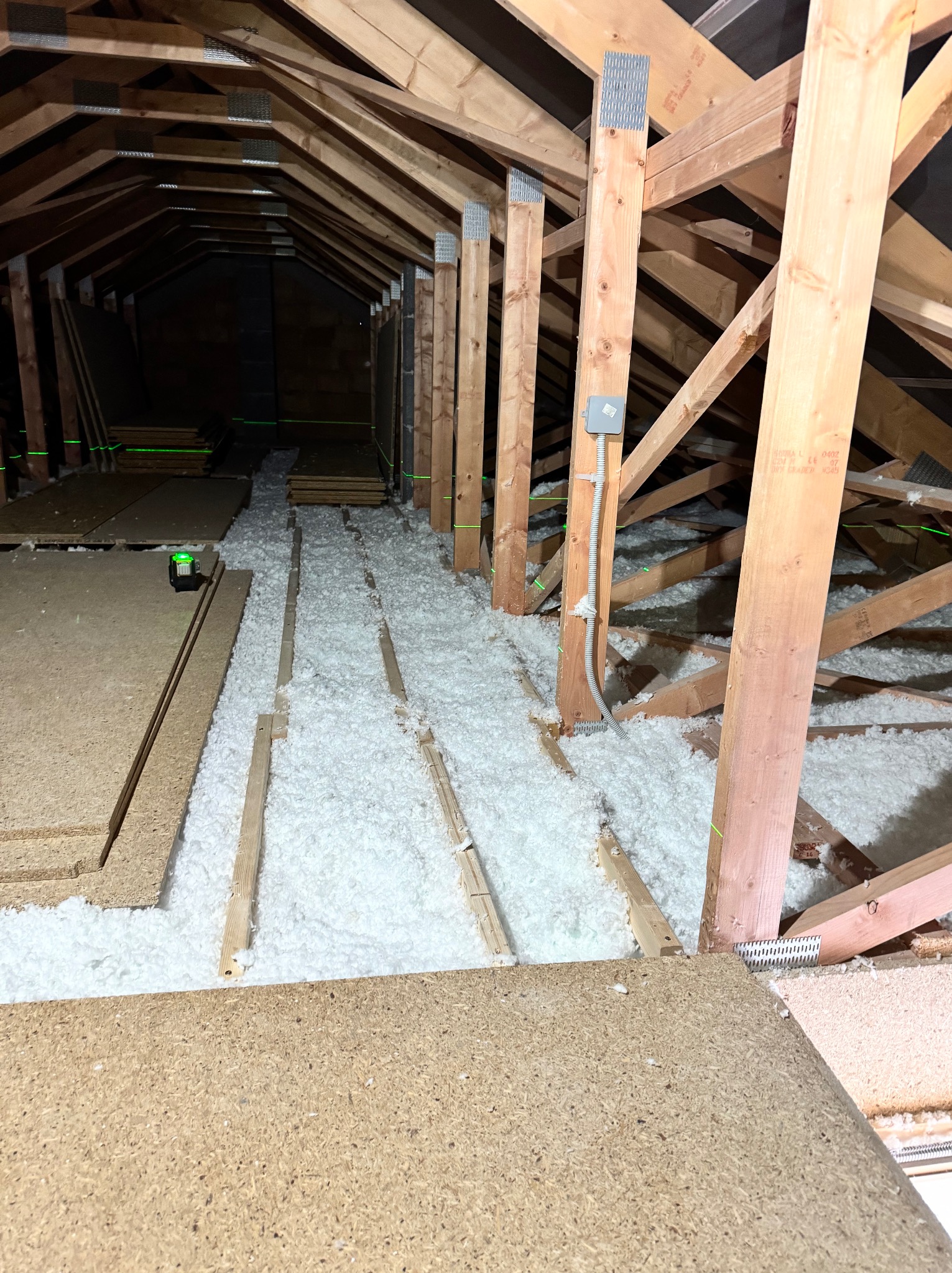 Poddasze w trakcie ocieplania: widoczna drewniana konstrukcja dachu, biała wełna izolacyjna między legarami, płyty OSB i poziomica laserowa.