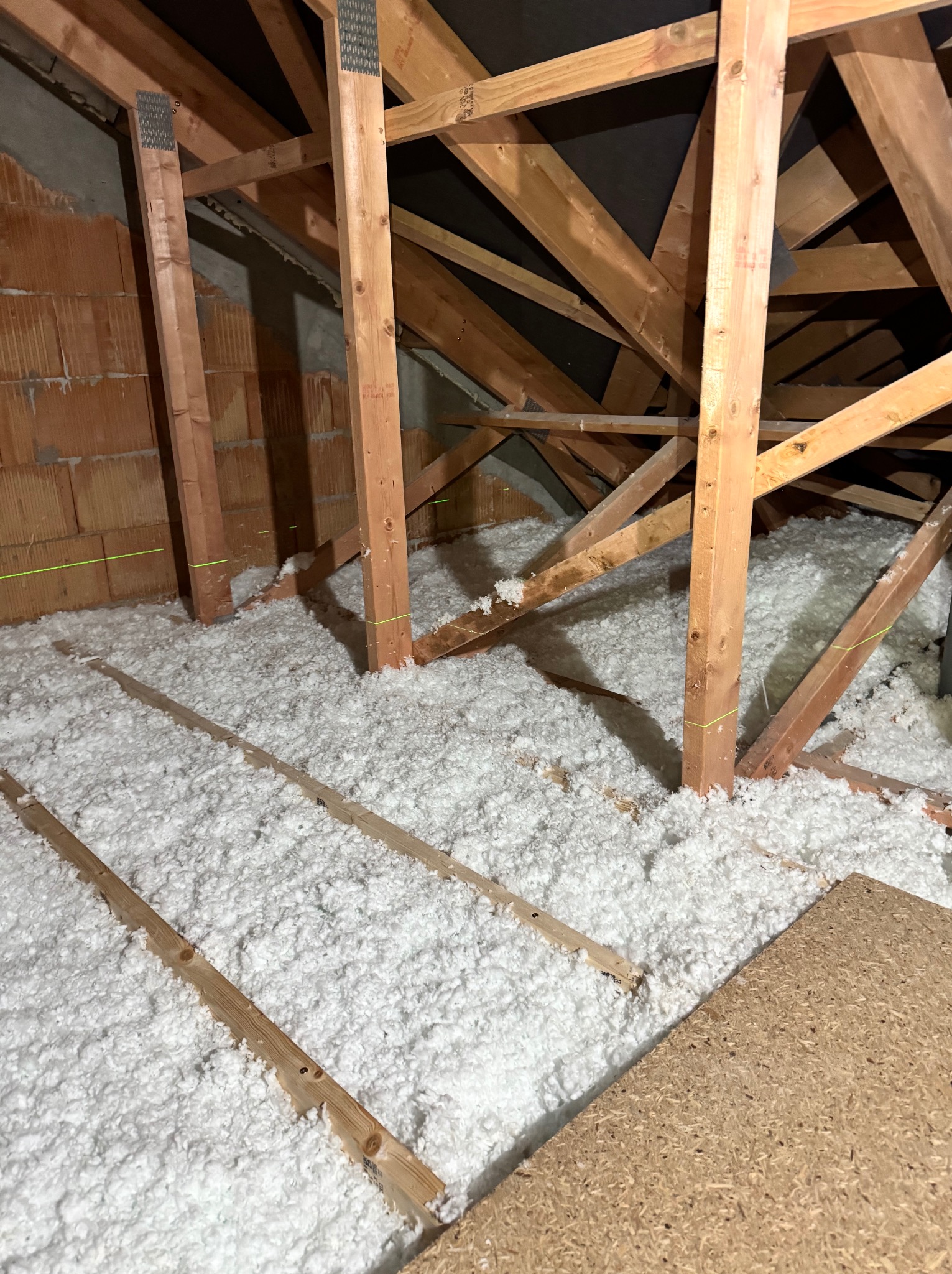 Poddasze w trakcie ocieplania wełną mineralną, widoczne krokwie drewniane, ceglany mur oraz poziome linie lasera wyznaczające poziom izolacji.