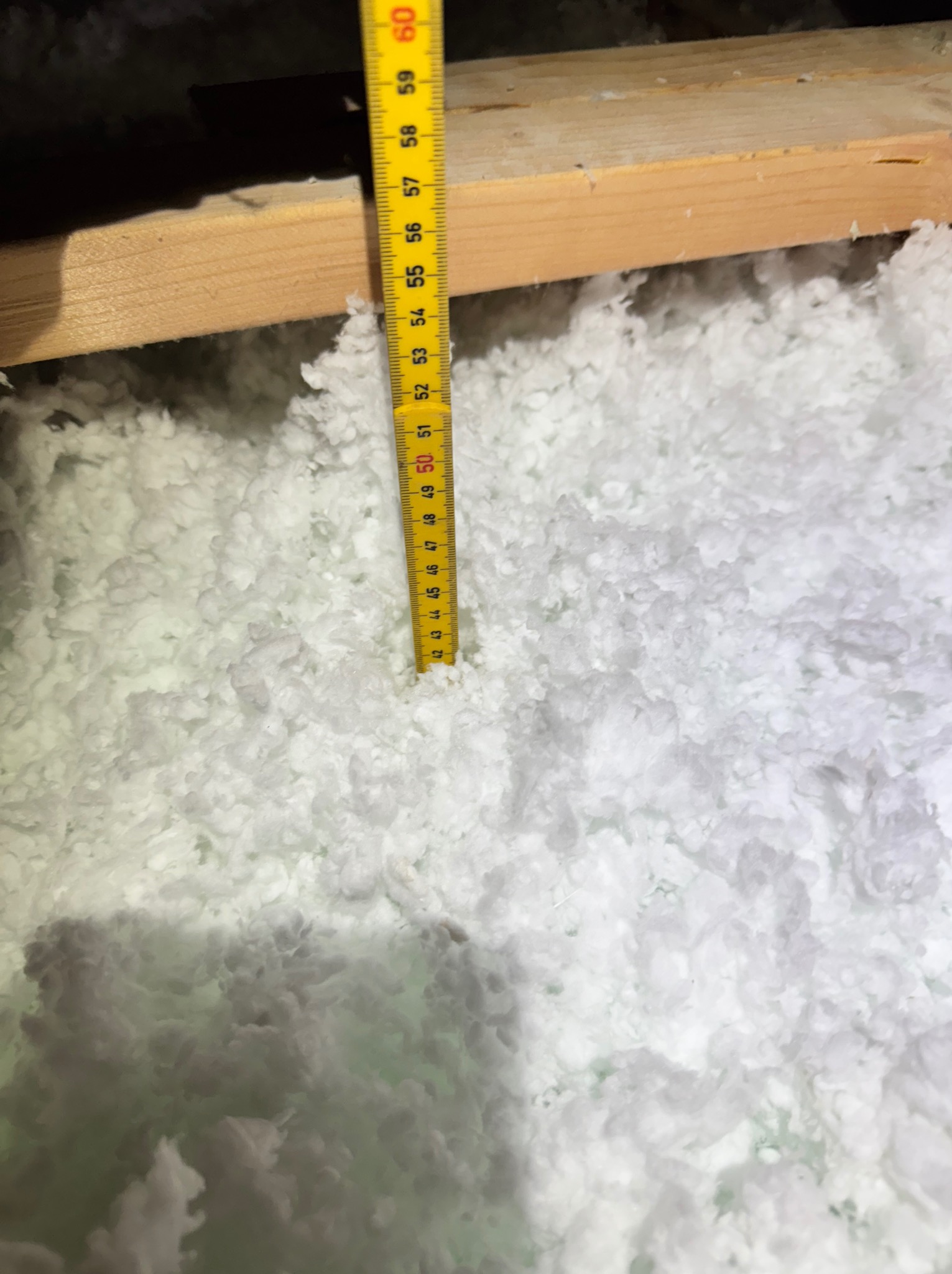 Pomiar grubości izolacji z białej wełny mineralnej w przestrzeni poddasza, widoczna drewniana belka i żółta miarka.