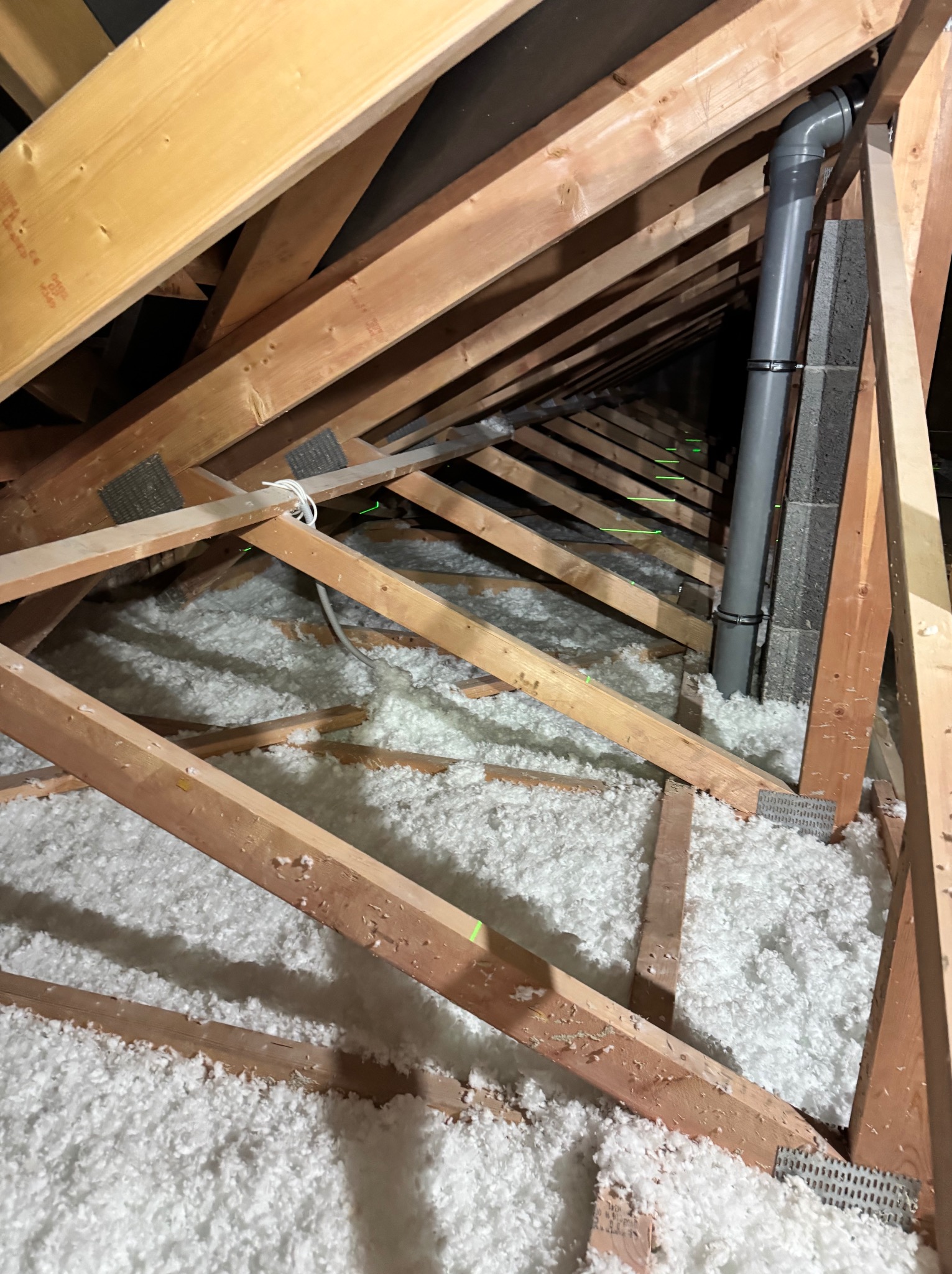 Poddasze z drewnianą konstrukcją dachu wypełnione białą izolacją z włókien, widoczny szary przewód wentylacyjny przymocowany do komina.