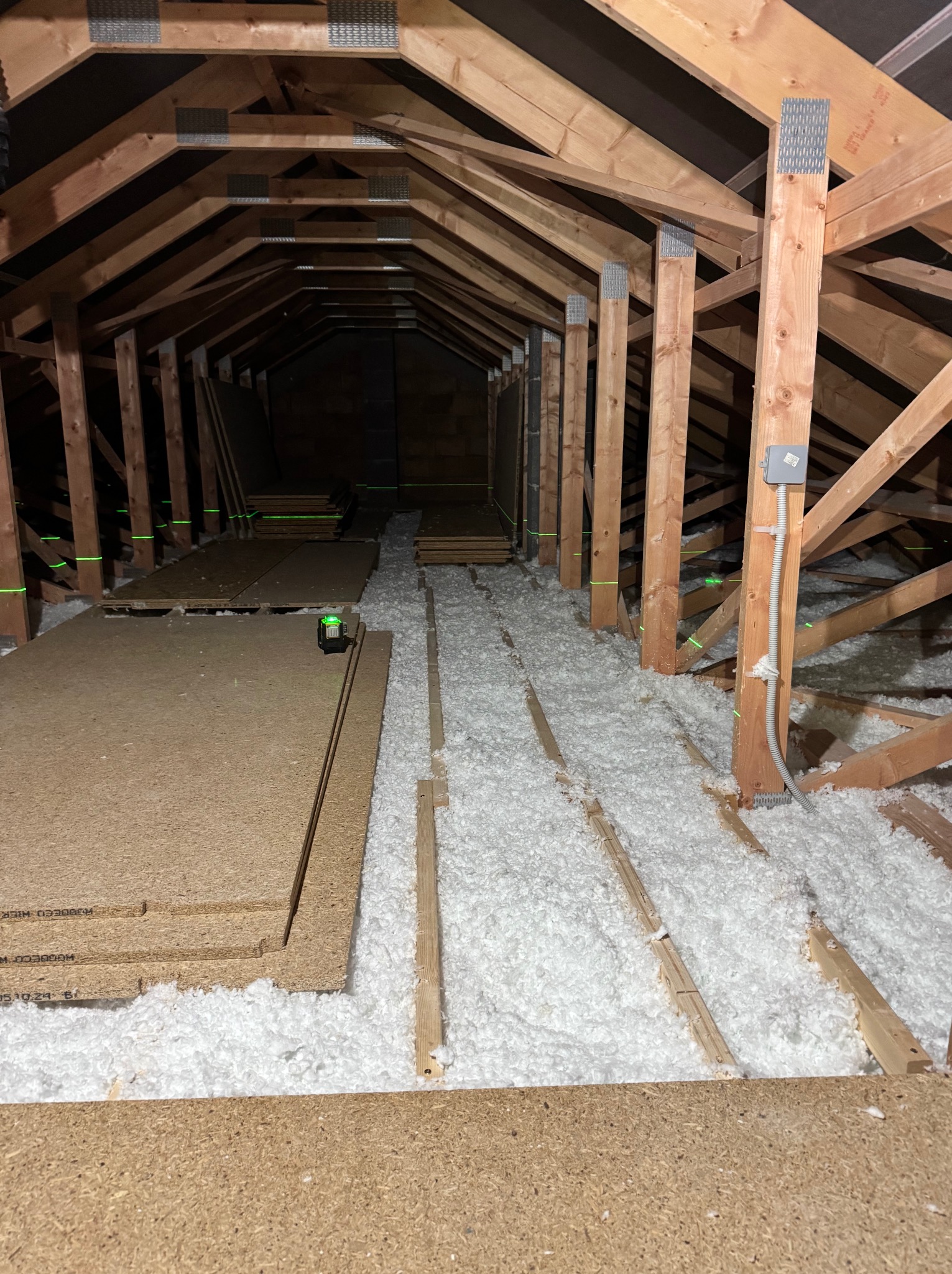 Poddasze w trakcie ocieplania: widoczne drewniane krokwie, biała wełna izolacyjna, płyty OSB i poziomica laserowa.