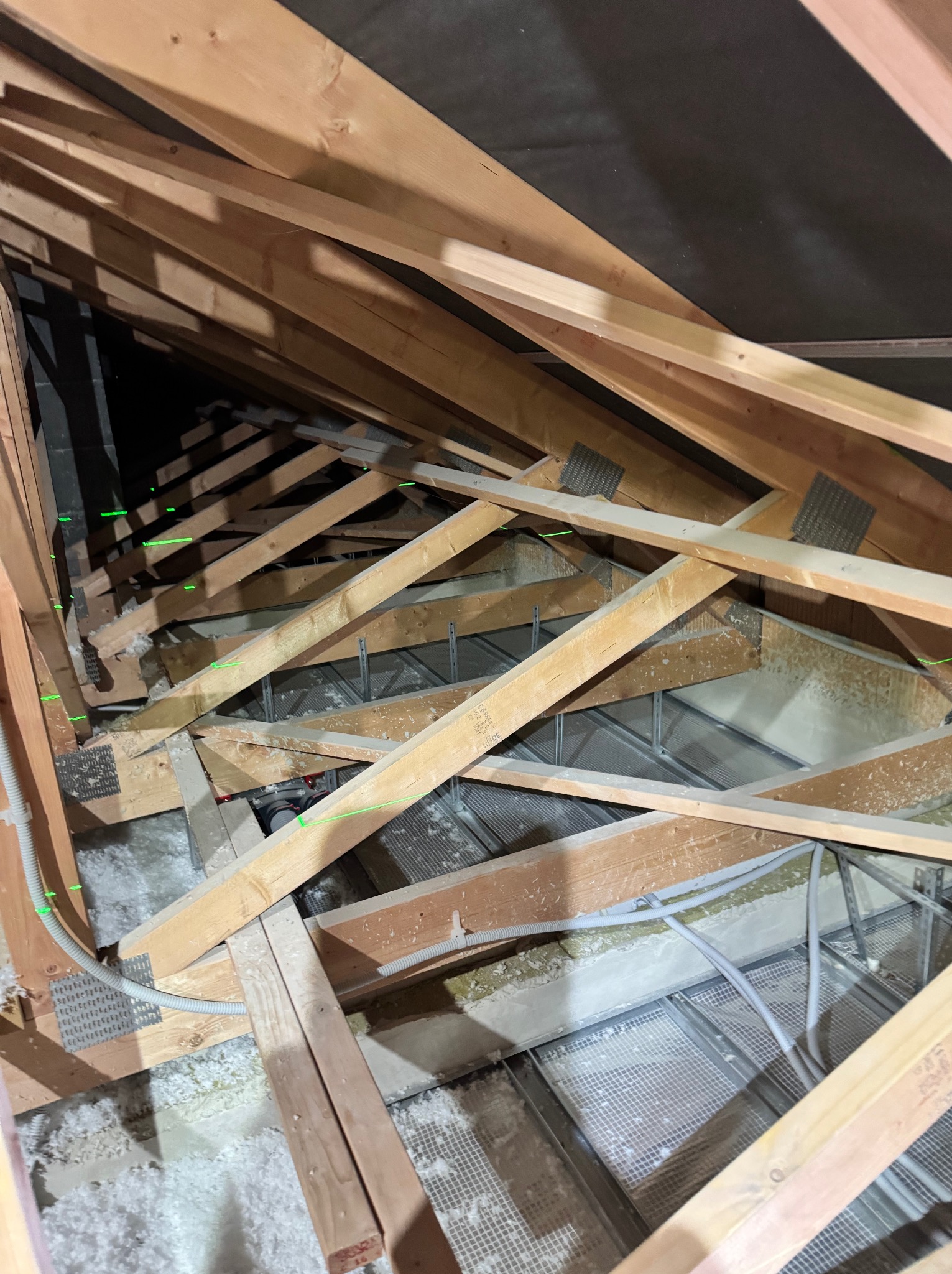 Szkielet drewniany poddasza z widoczną izolacją z wełny mineralnej, metalową siatką i przewodami elektrycznymi, oznaczony zielonym laserem poziomującym.