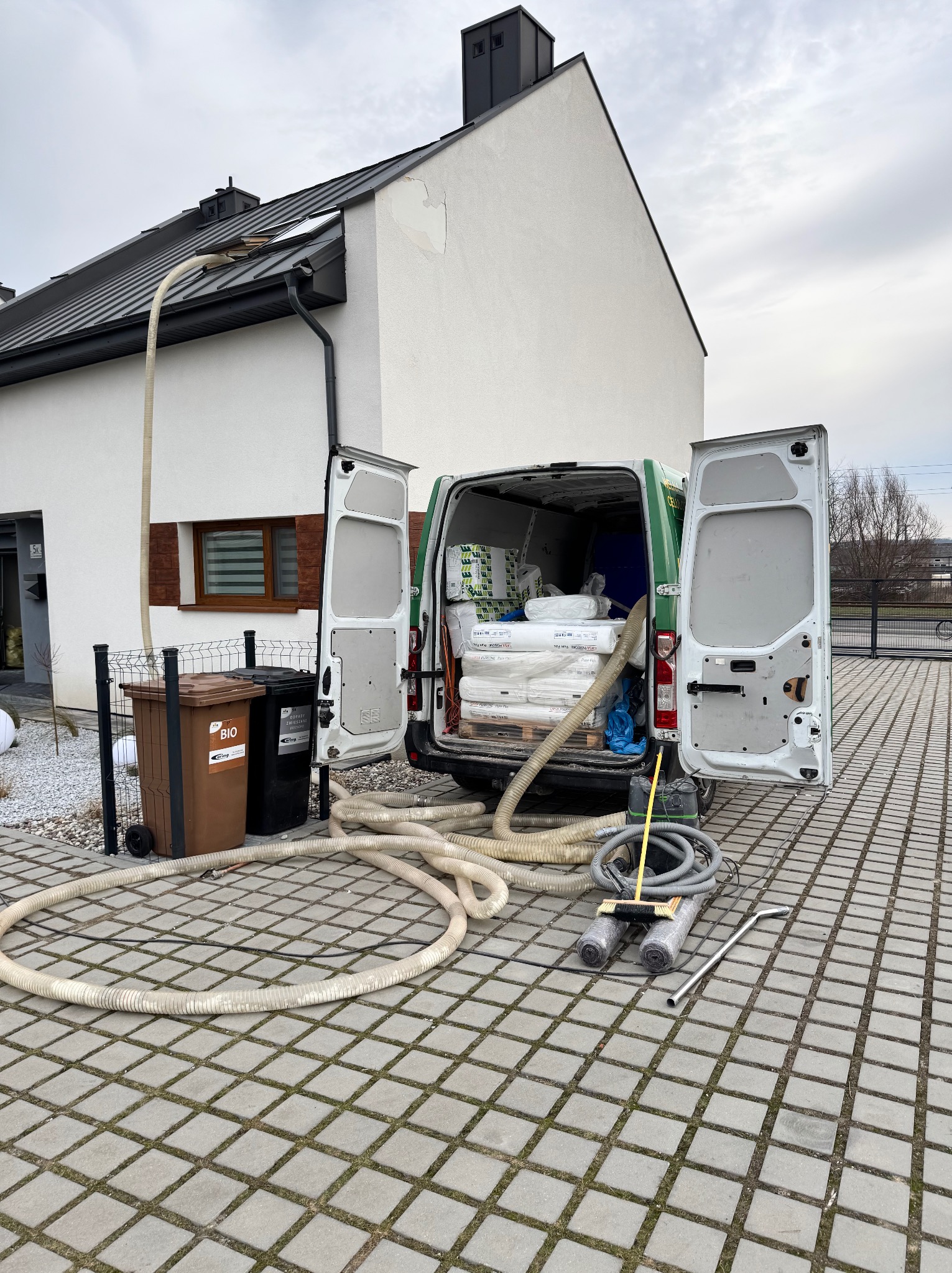 Samochód dostawczy z otwartymi drzwiami, wypełniony materiałami izolacyjnymi, z podłączonym wężem do ocieplania poddasza, stojący na kostce brukowej przed domem z białą elewacją.
